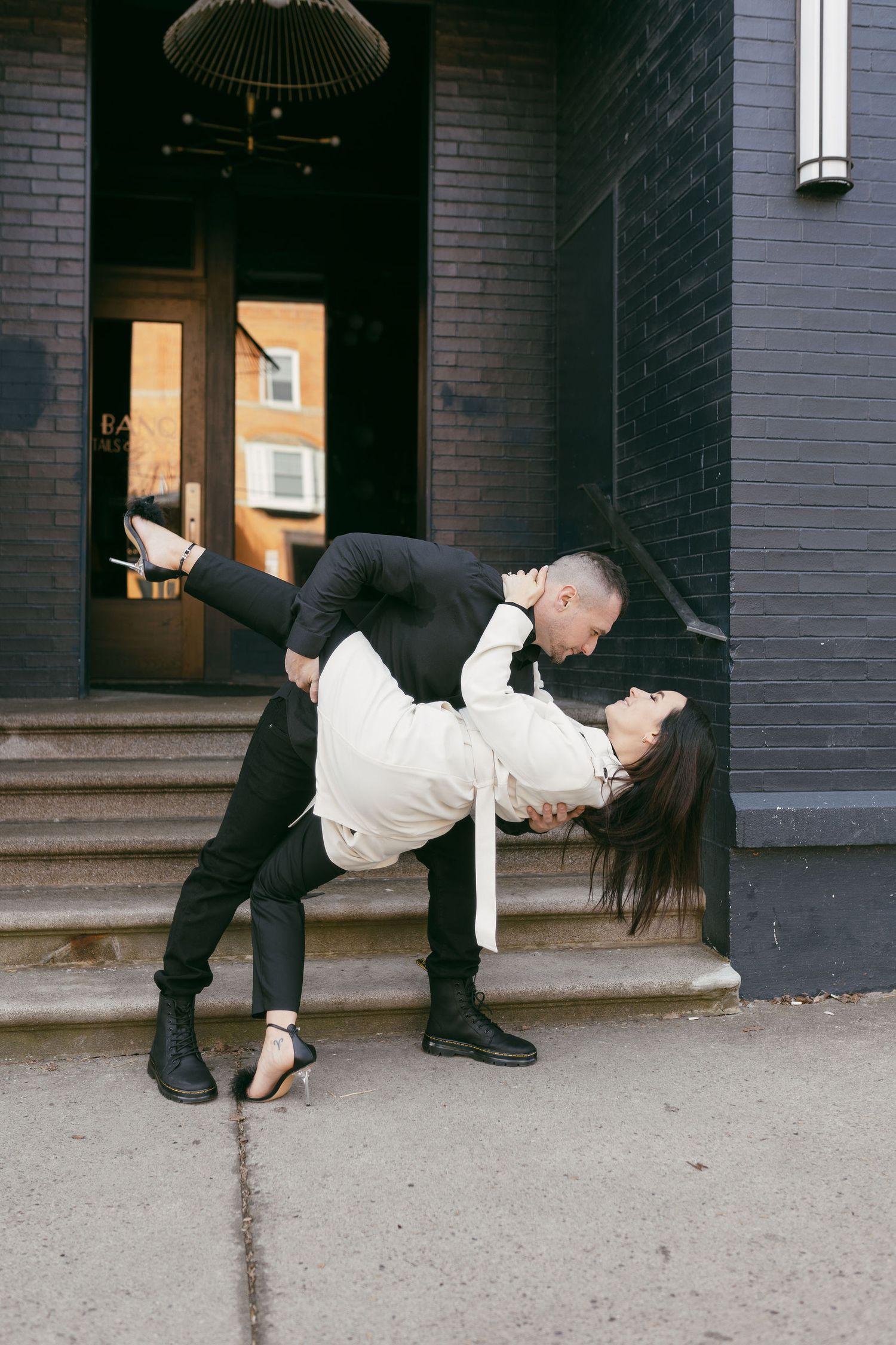 Couple sharing a romantic dip kiss outside a dark brick building on a city sidewalk.