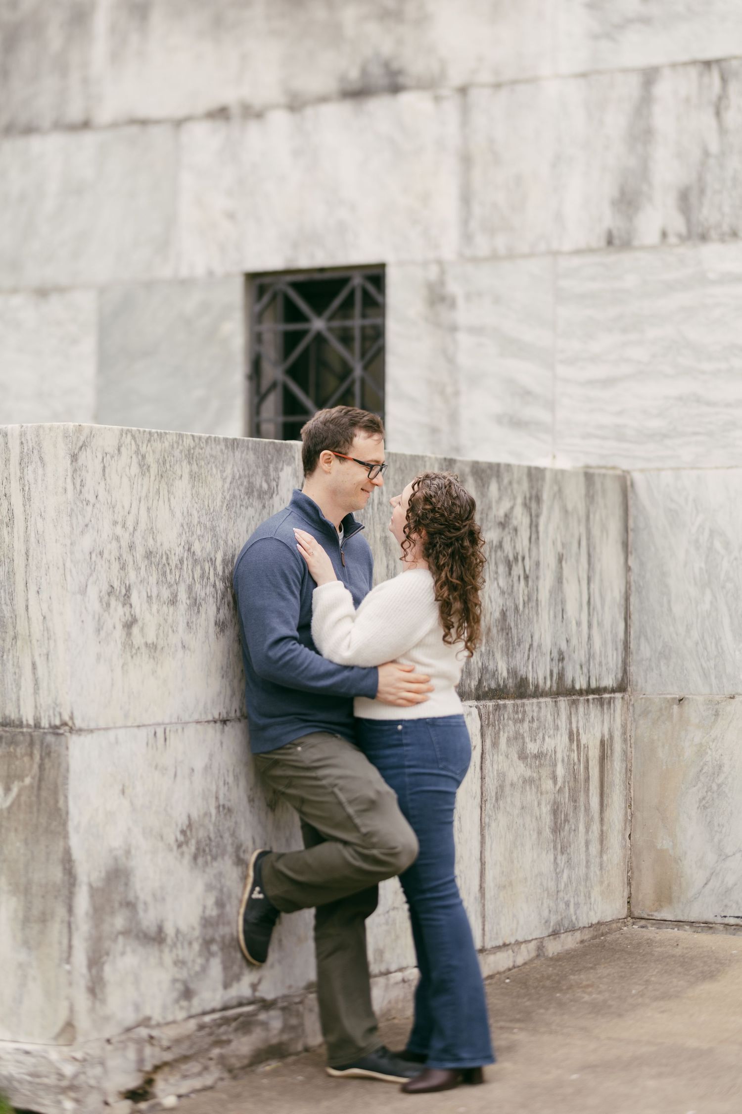 Couple embracing and smiling at each other against a marble wall outdoors, man in blue sweater, woman in jeans.
