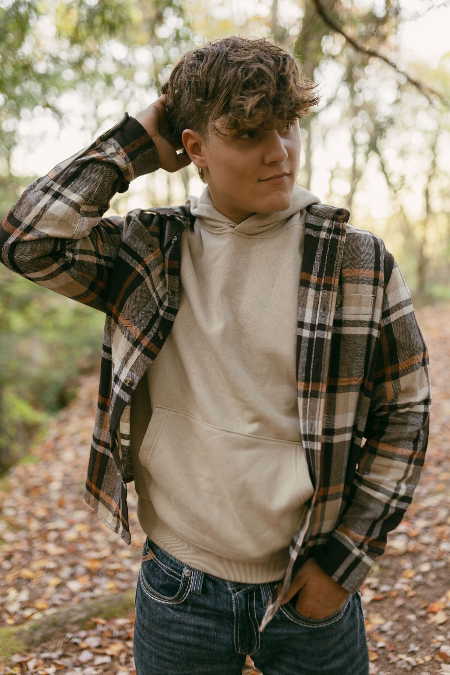 Young man in plaid flannel shirt and beige hoodie posing in autumn forest.