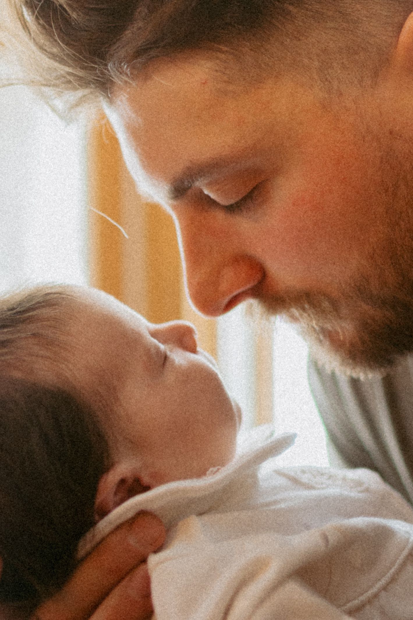 A bearded father tenderly touches noses with his sleeping newborn baby, sharing a gentle and loving moment.