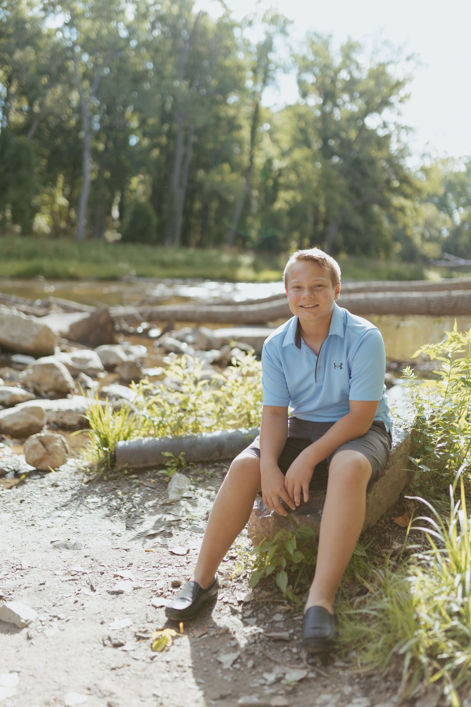 A smiling teenage boy in a light blue polo shirt sits on a log in a natural outdoor setting with trees and rocks.