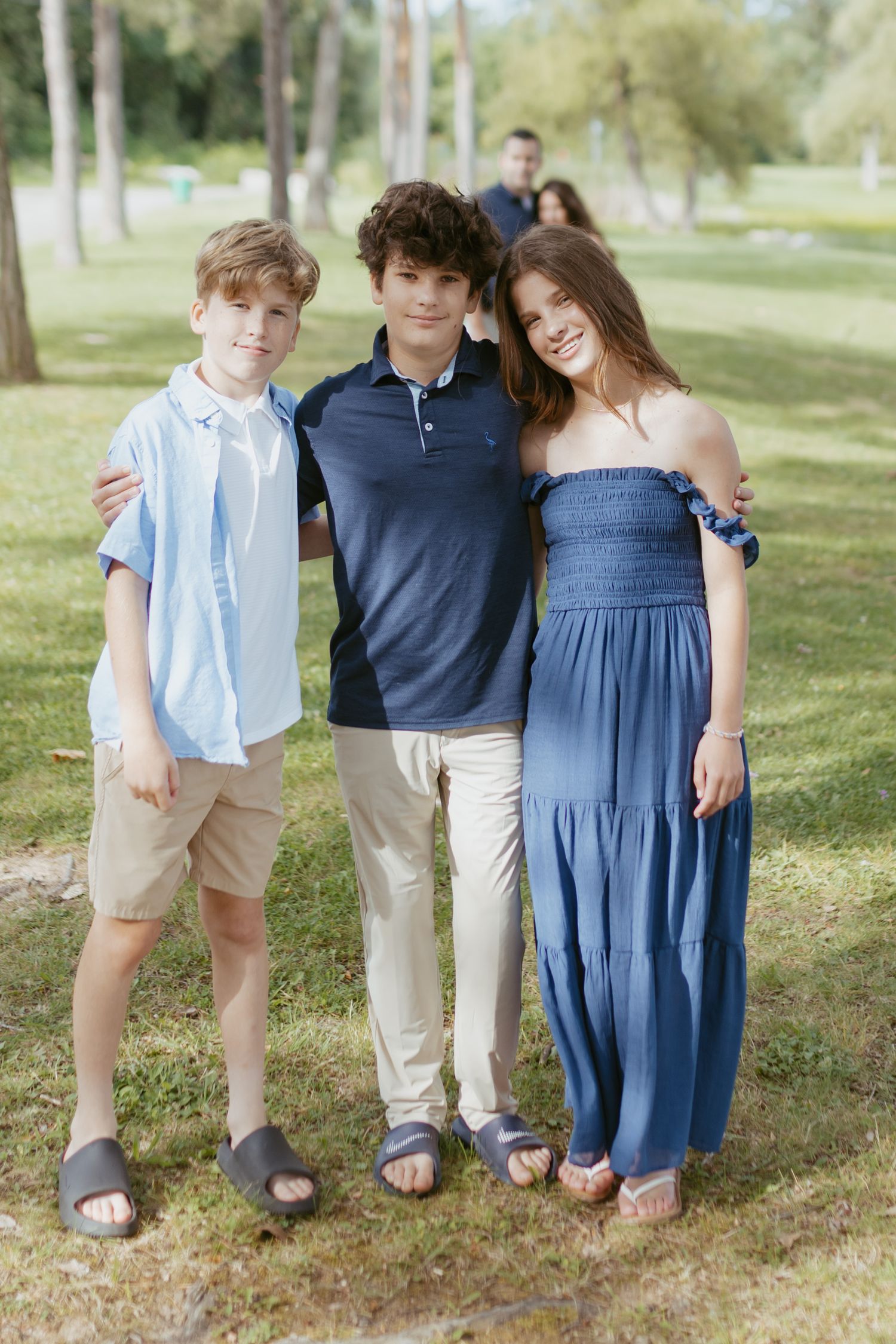 Three teenagers posing together outdoors in a green park, wearing blue outfits, smiling for a family photo.