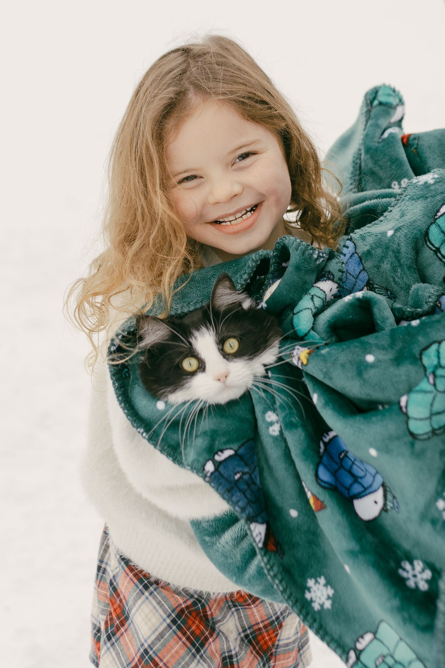 Smiling girl with curly hair holds black and white cat in teal snowflake blanket outdoors in snow.