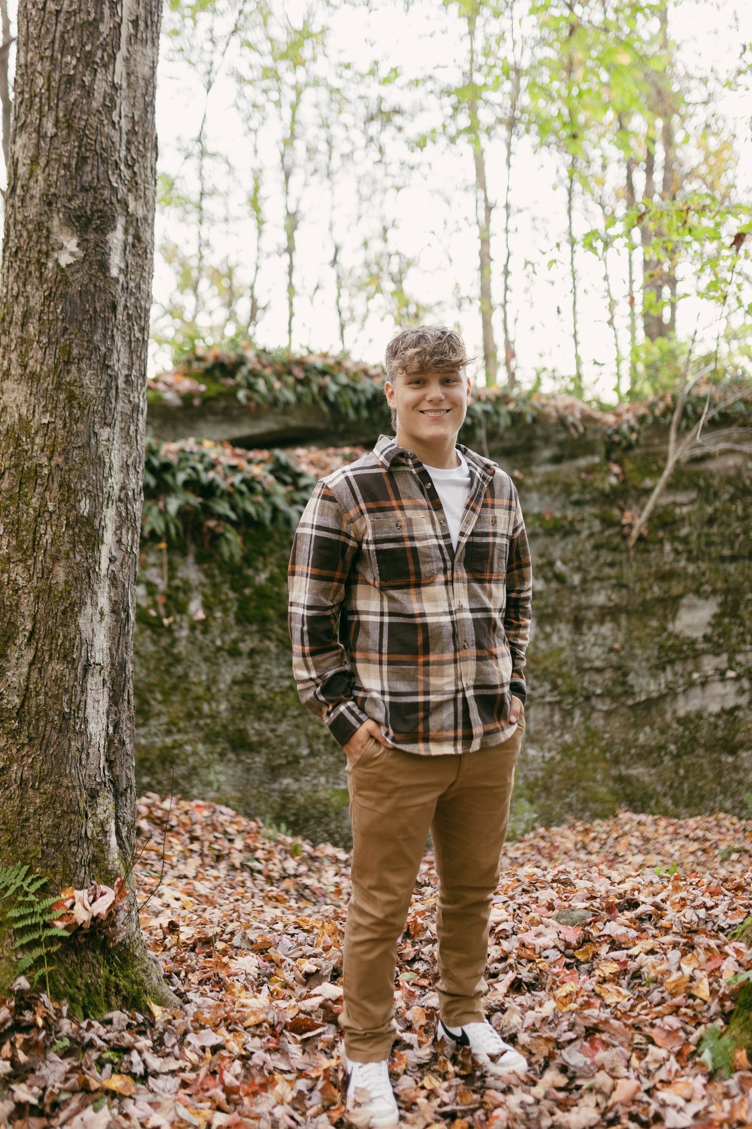 Young man in plaid shirt and tan pants smiling among autumn leaves in a wooded forest setting.