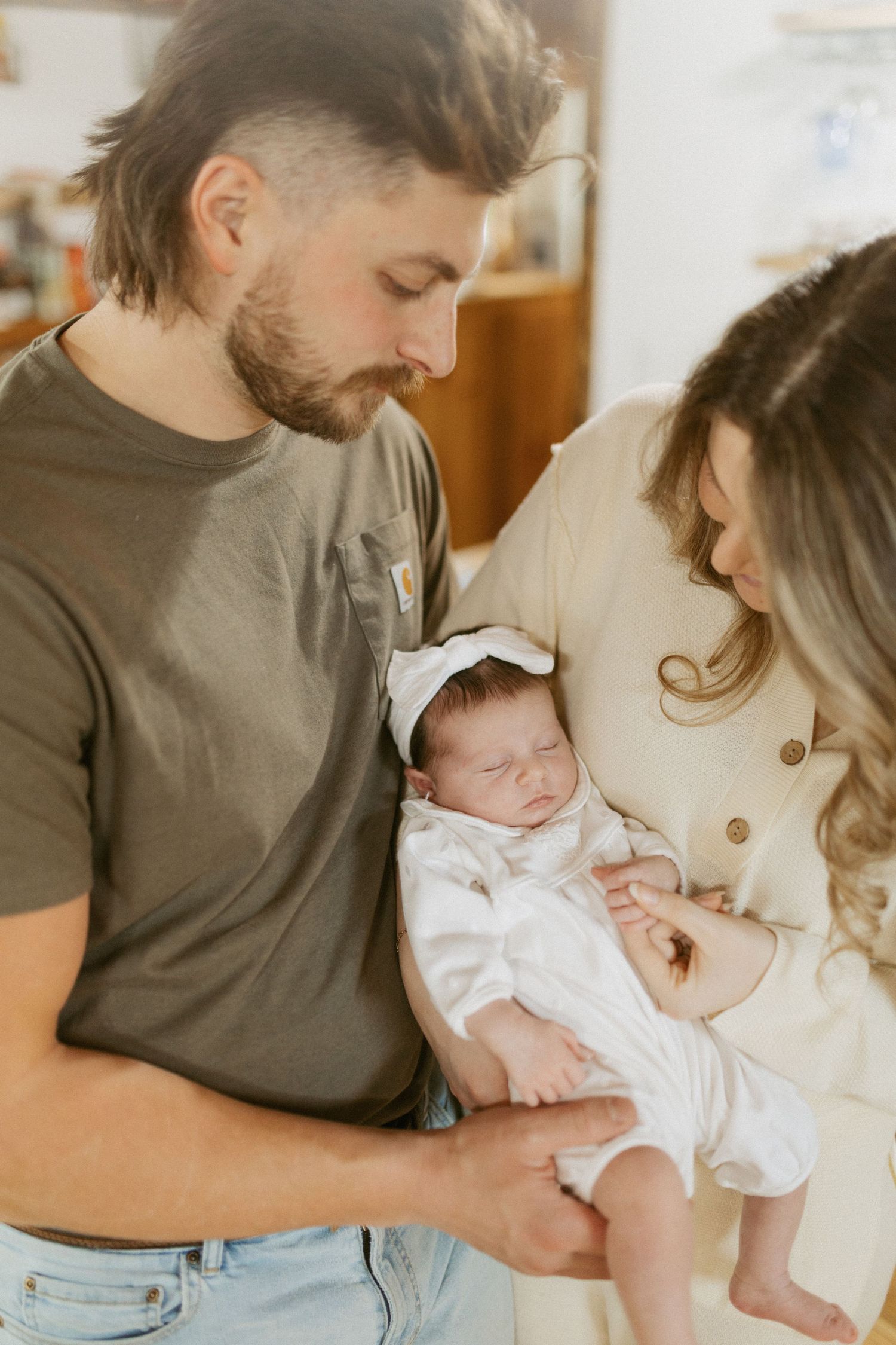 New parents lovingly gaze at their sleeping newborn baby dressed in white with a bow headband.