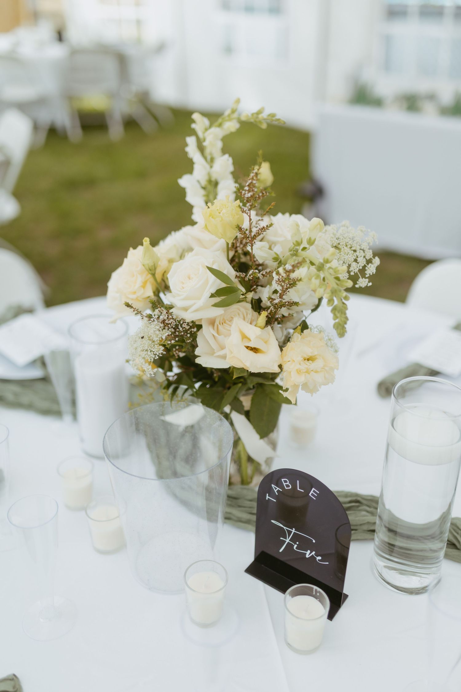 Elegant wedding reception table five with white floral centerpiece, candles, and glass vases on white linen.