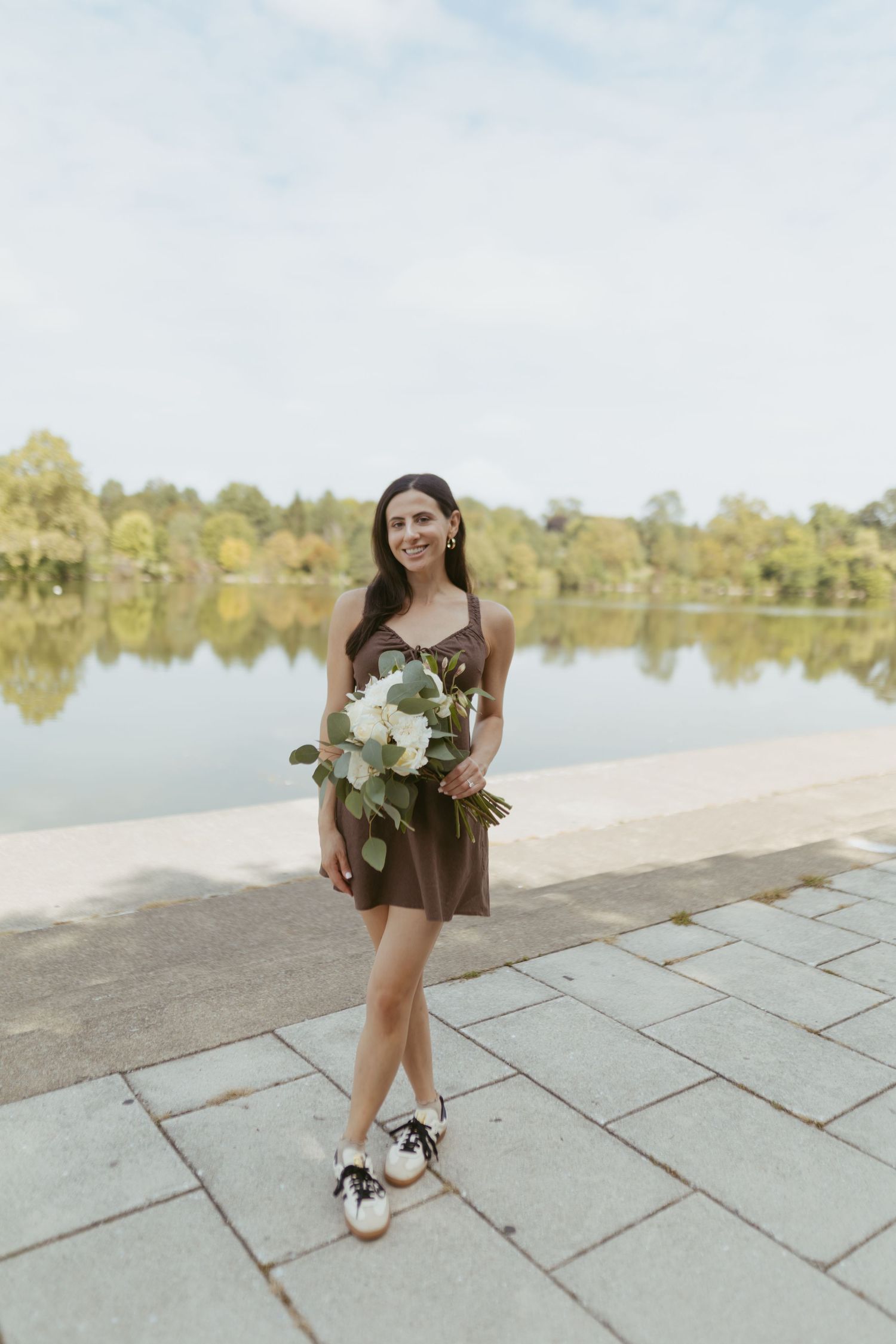 Smiling woman in brown dress holding white flower bouquet standing by a reflective lake in a park.