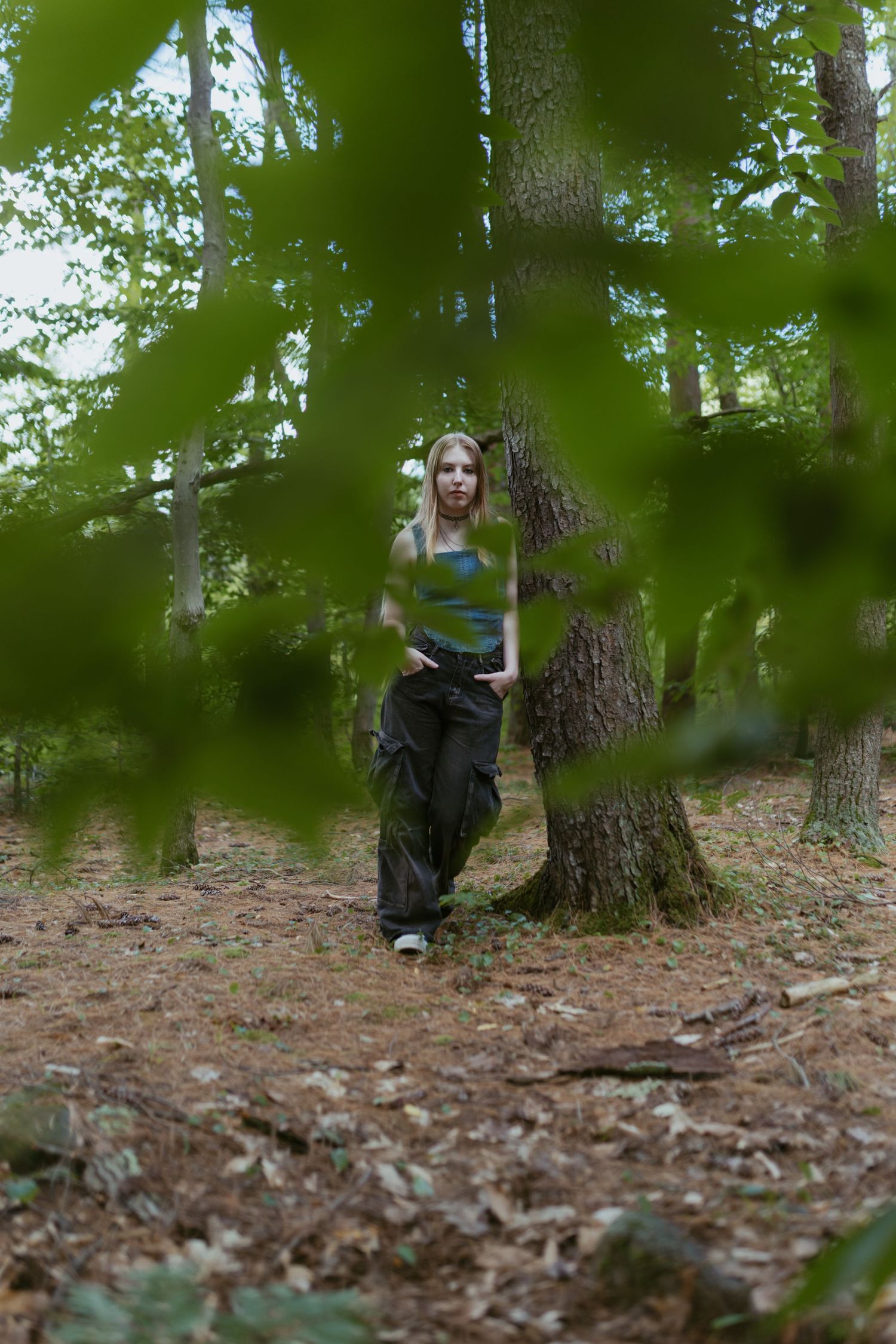 A young woman in a green tank top and cargo pants poses beside a tree in a lush green forest.