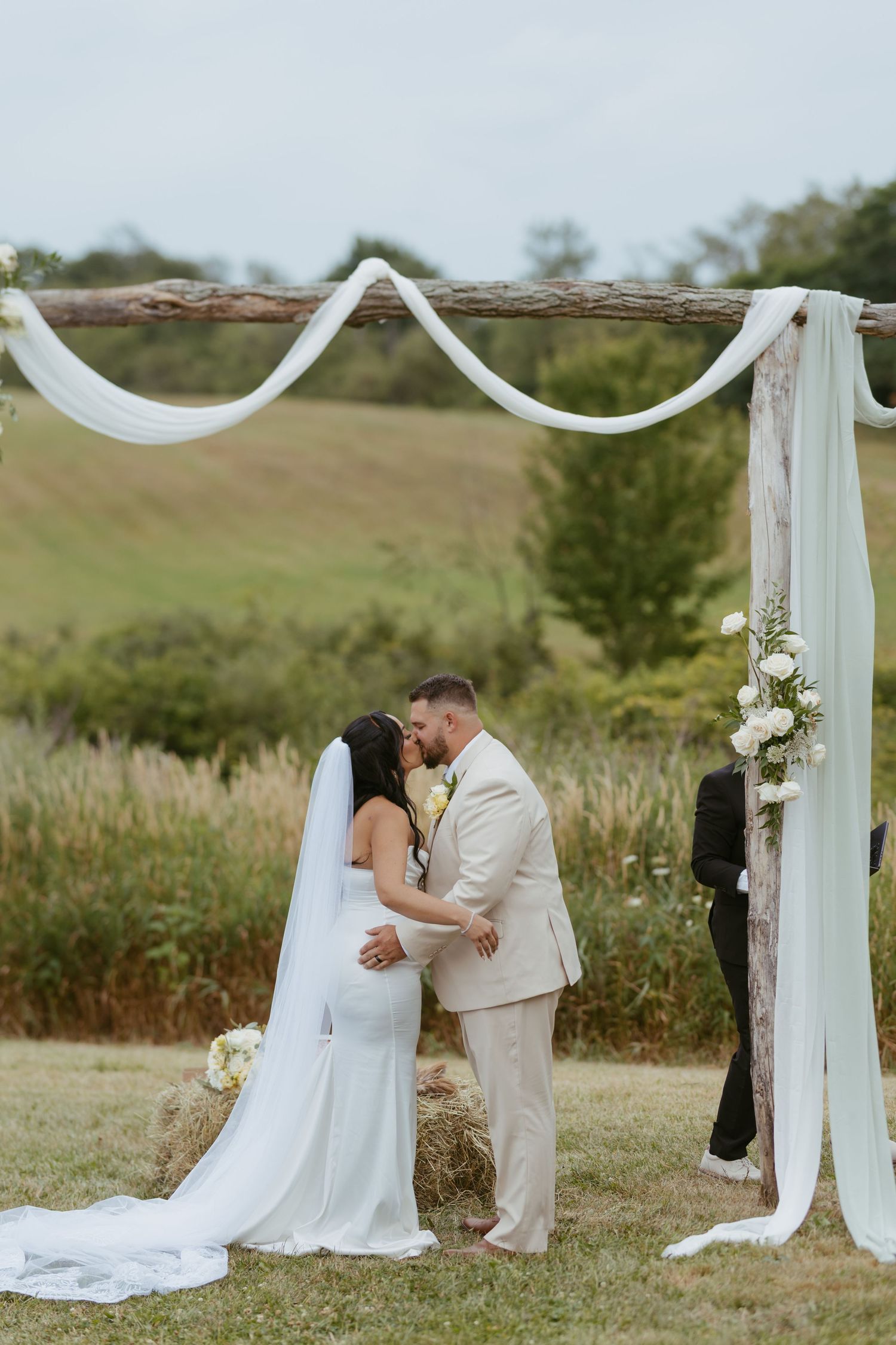 Bride and groom share first kiss under rustic wooden arch with white draping at outdoor farm wedding ceremony.