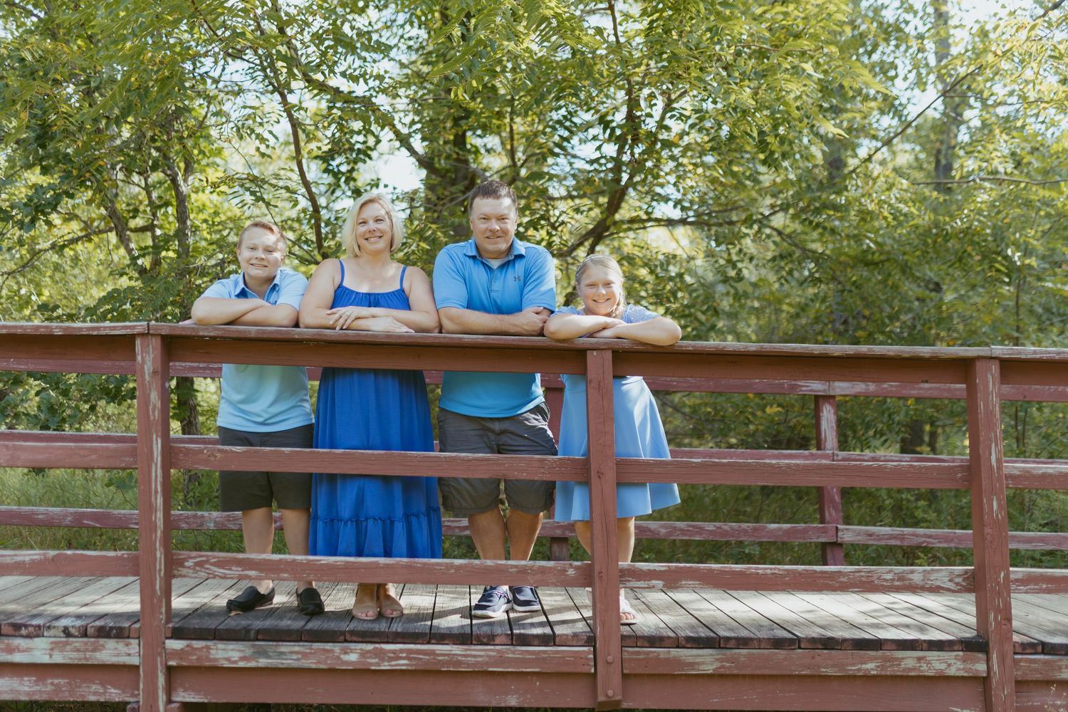 Family of four posing on a wooden bridge surrounded by lush green trees on a sunny day.