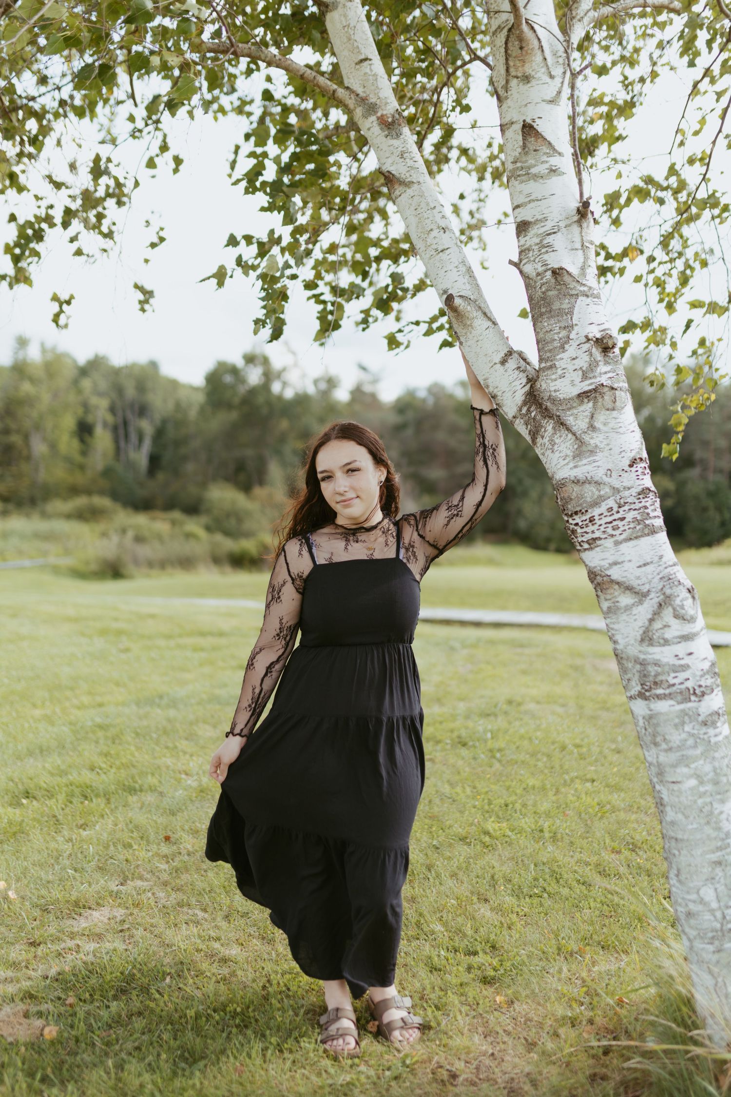 Young woman in black lace dress posing by a birch tree in a green outdoor park setting.