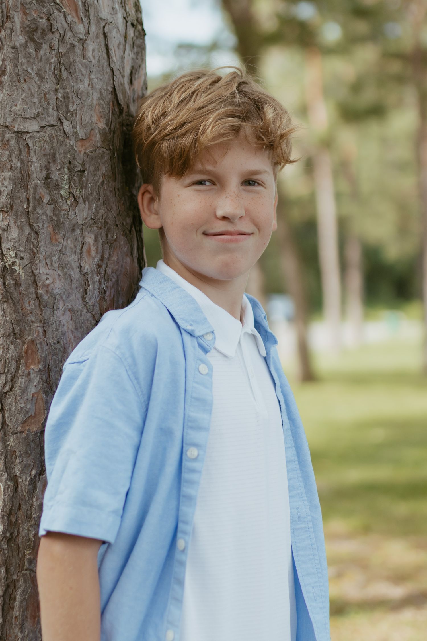 A smiling teenage boy with blonde hair leans against a tree trunk in a sunny park, wearing a light blue shirt.