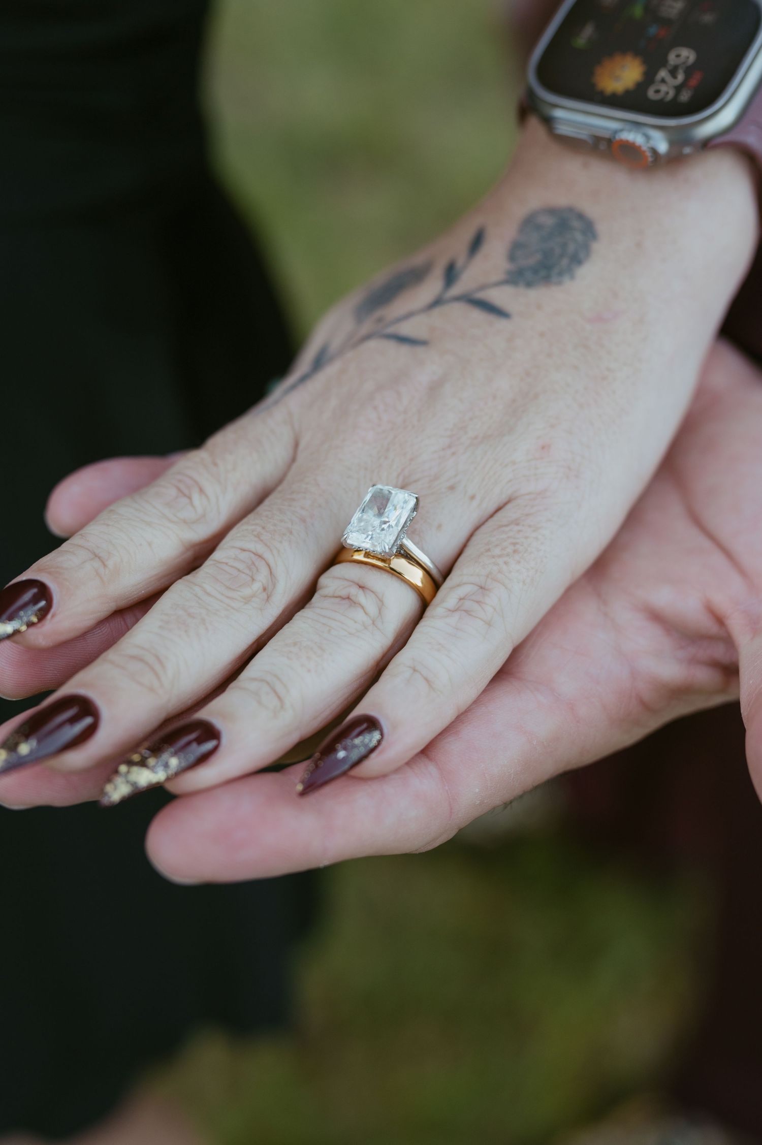 Close-up of hands displaying a gold engagement ring with emerald-cut diamond, dark manicure, and floral tattoo.