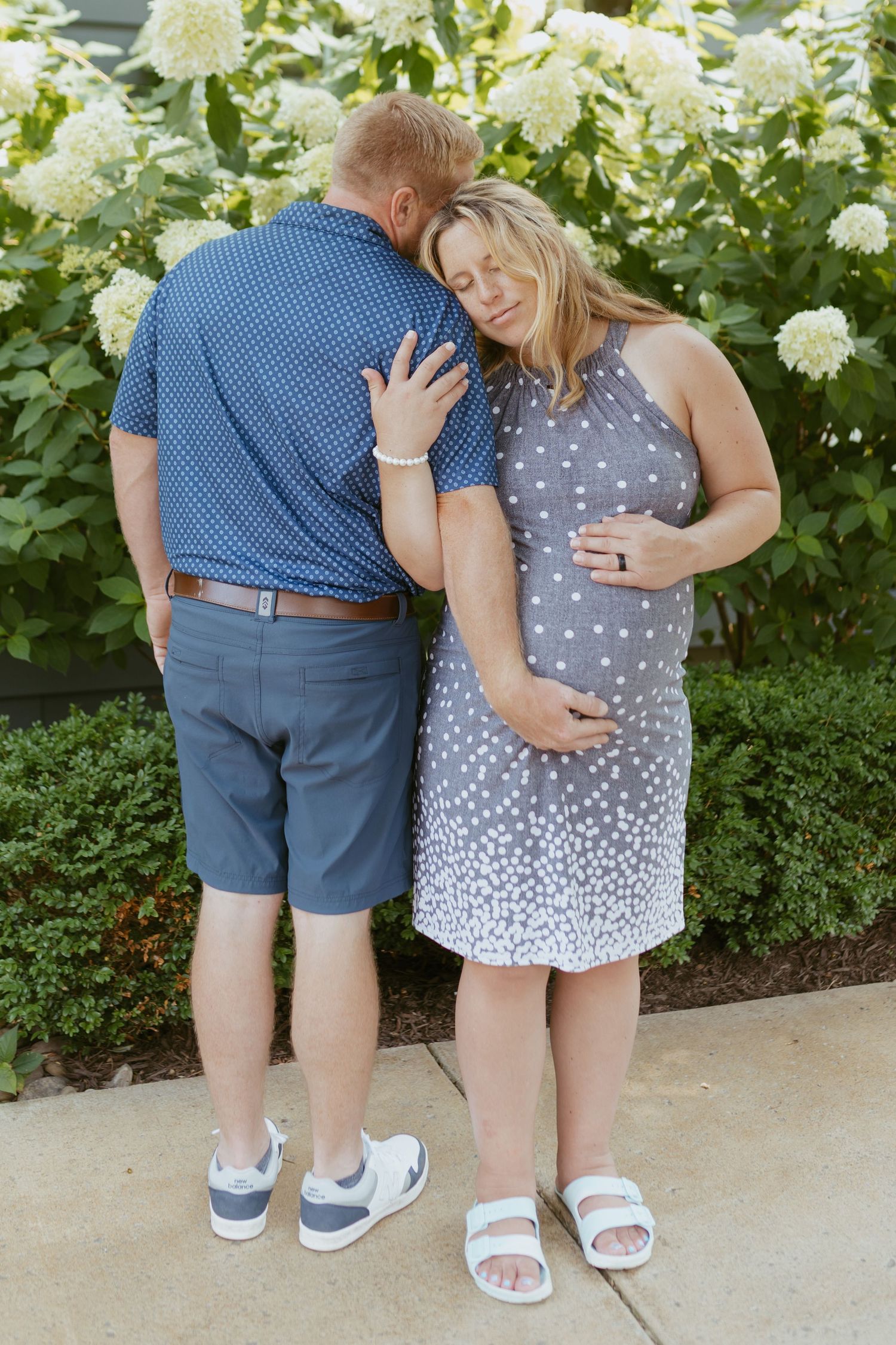 Pregnant woman in gray polka dot dress embraces man in blue shirt outdoors near white hydrangea bushes.