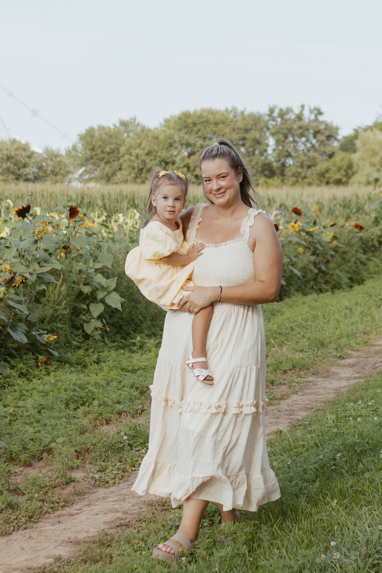 A smiling woman in a cream dress holds a young girl in yellow near a sunflower field outdoors.