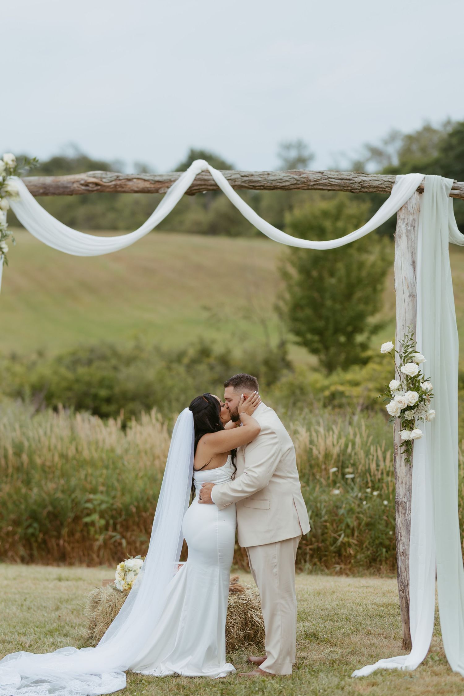 Bride and groom share first kiss under rustic wooden arch with white draping and floral accents at outdoor wedding ceremony.
