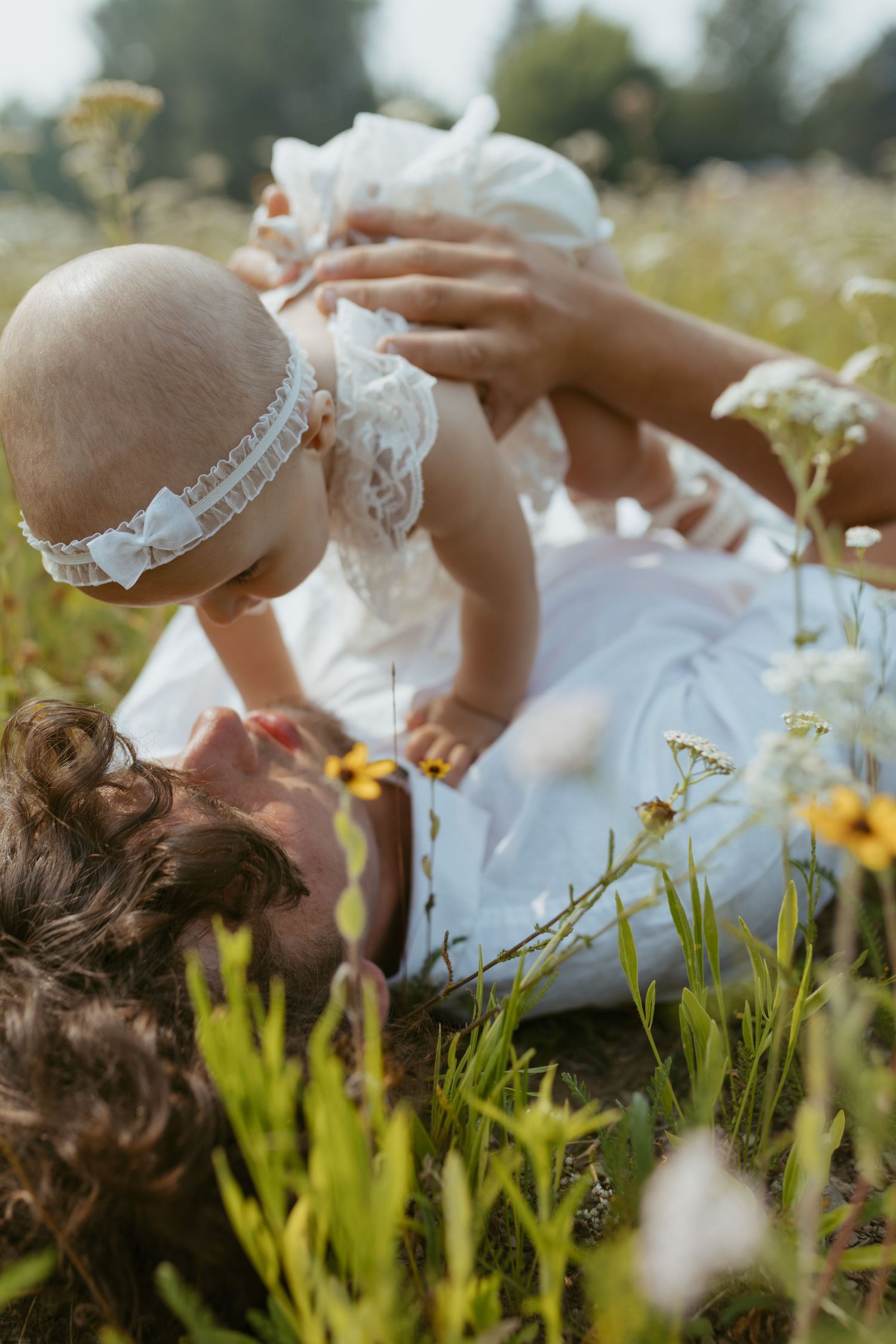 Mother lying in wildflower field lifting smiling baby dressed in white lace on sunny day.