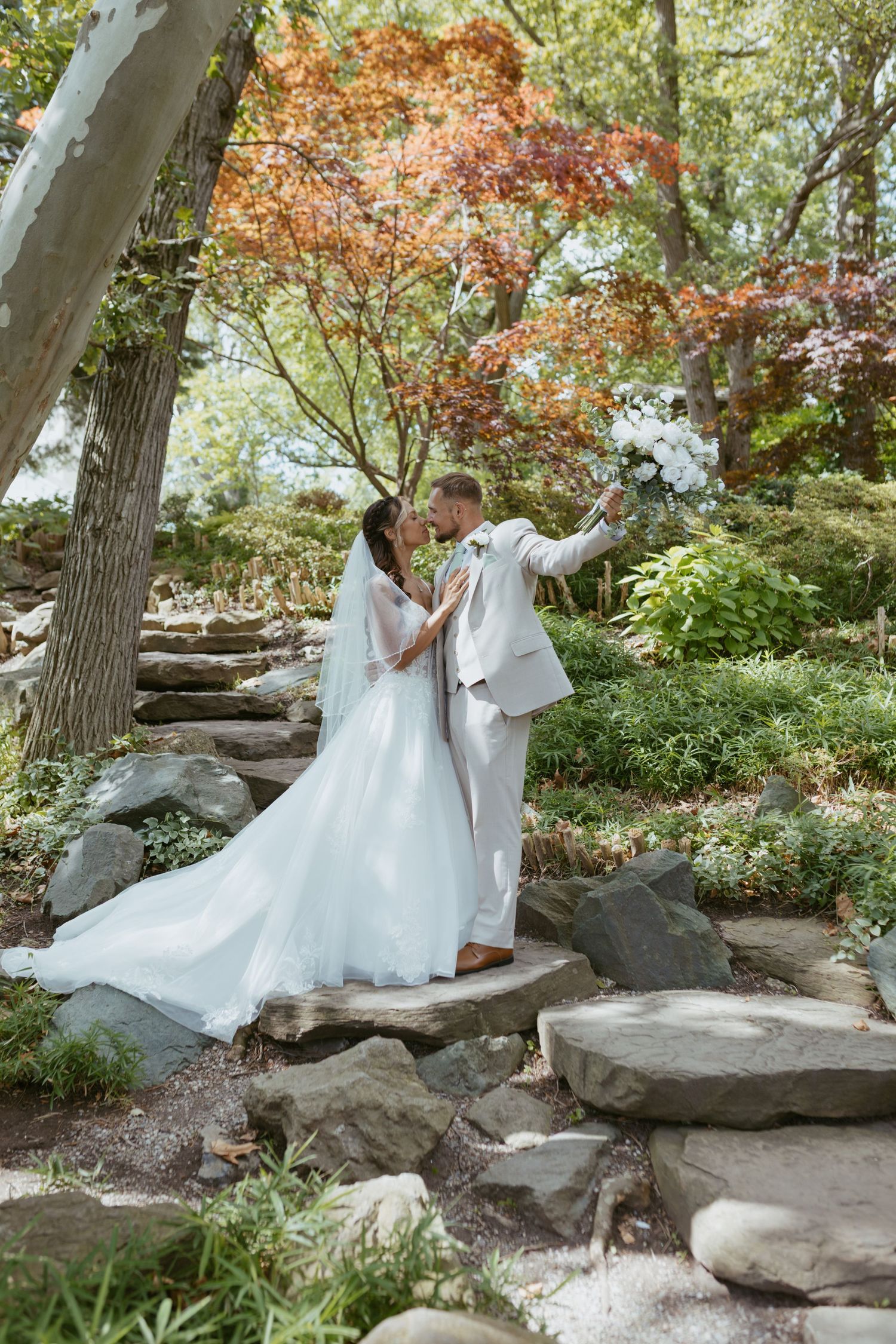 A bride and groom share a romantic moment on stone steps surrounded by lush greenery and autumn trees in a garden.