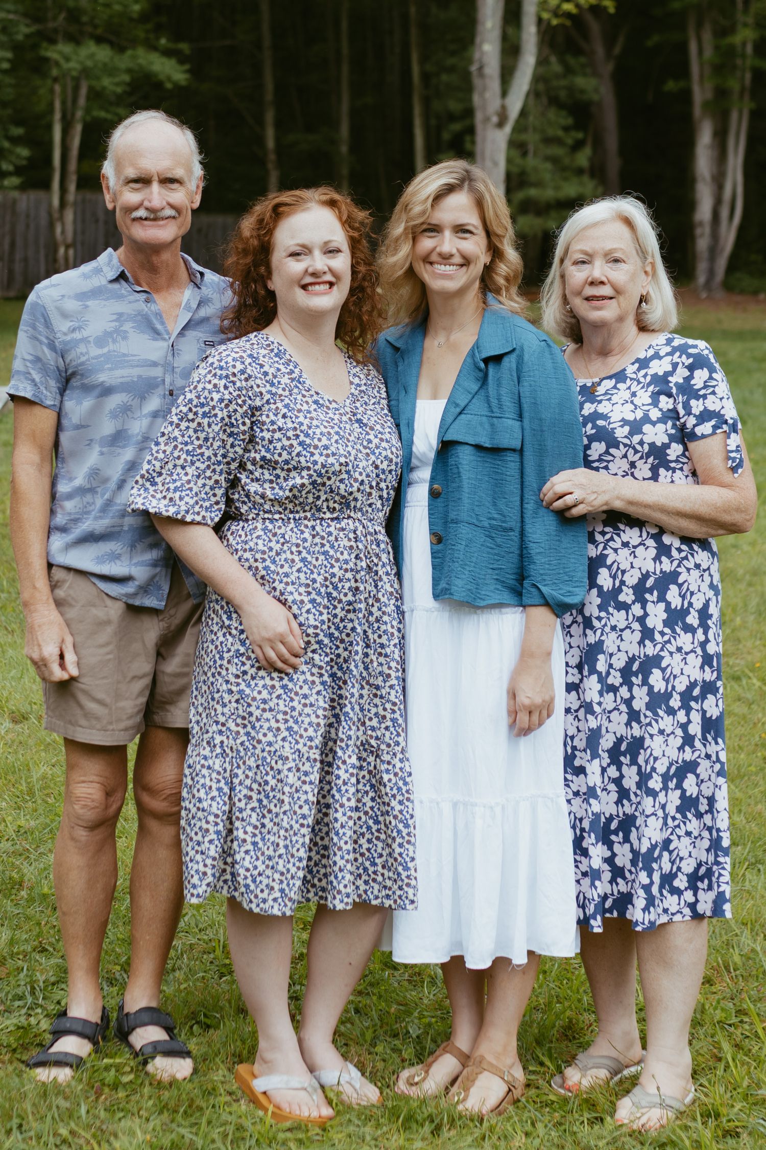 Family of four smiling outdoors in summer attire, posing together on green grass with trees in the background.