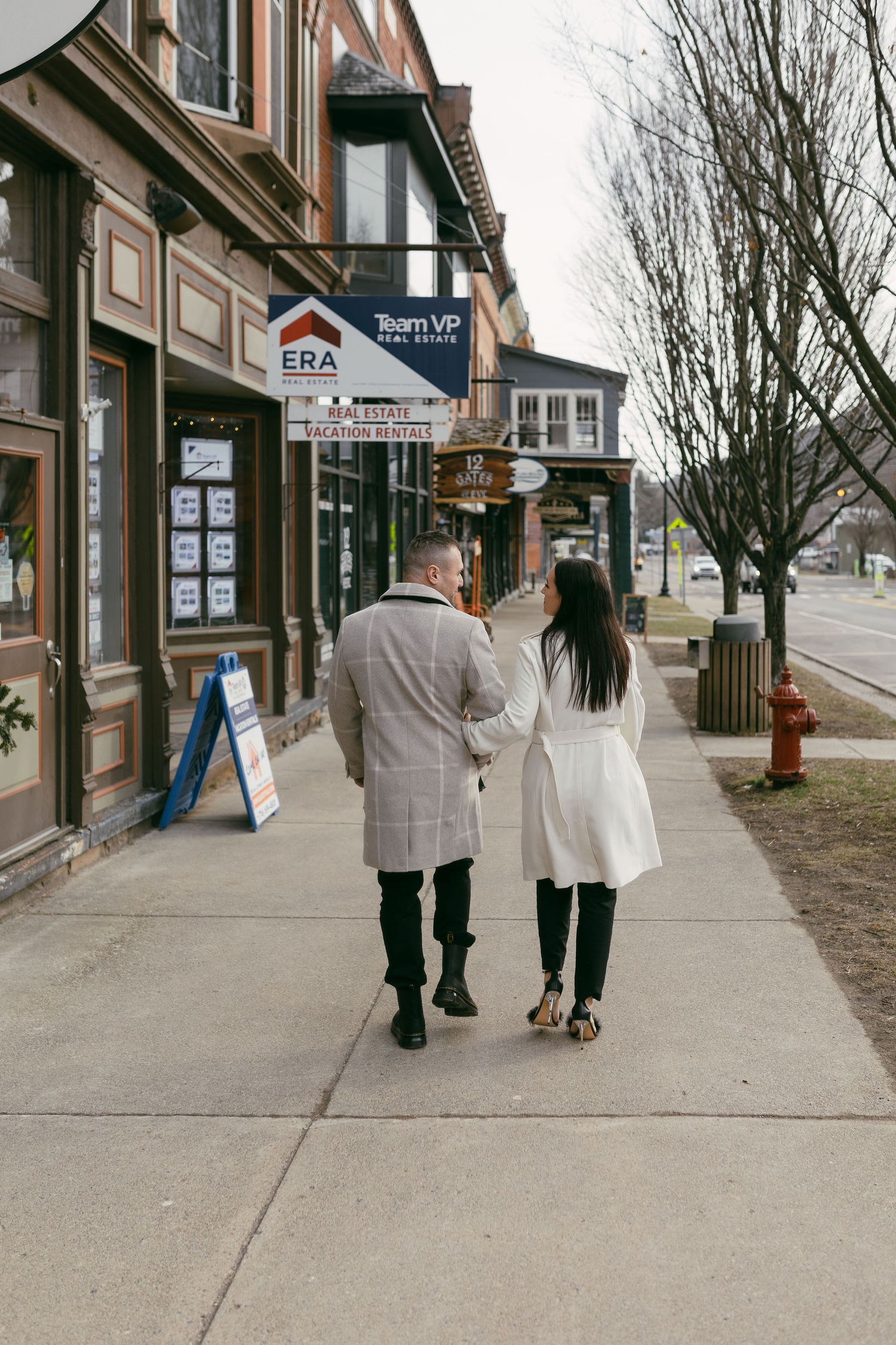 Couple walking hand-in-hand along a charming small-town sidewalk, wearing matching white and plaid coats in winter.