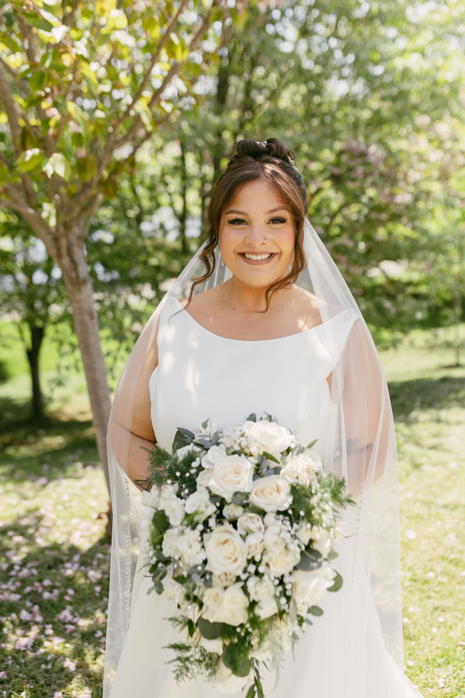 Smiling bride in white gown and veil holding large white rose and greenery cascading bouquet outdoors.