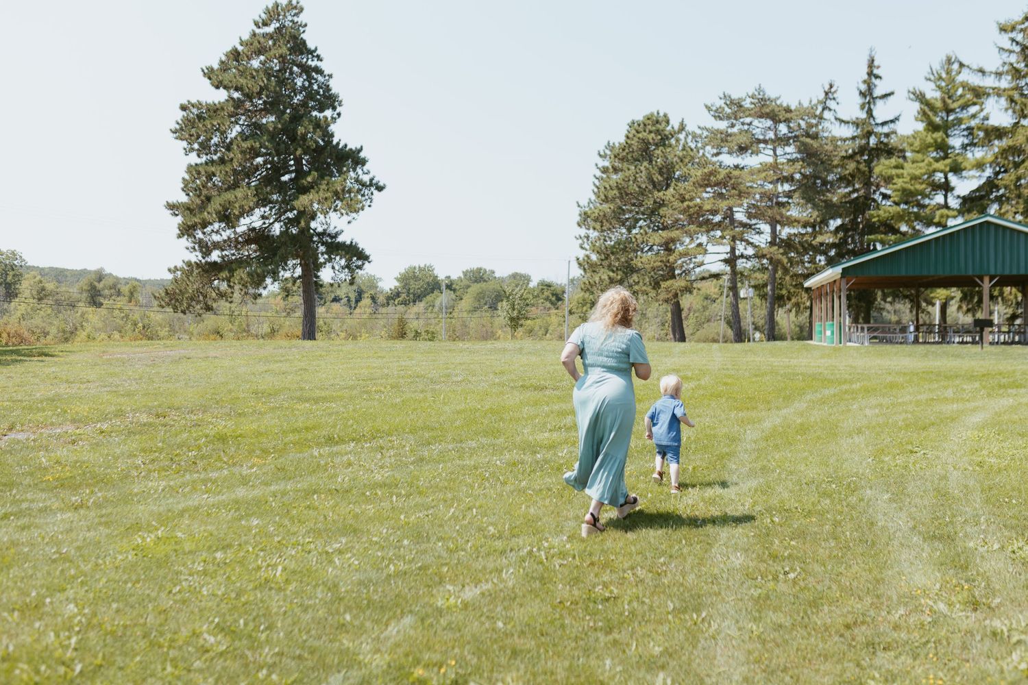 A woman in a teal dress walks hand-in-hand with a toddler across a sunny green field with trees.