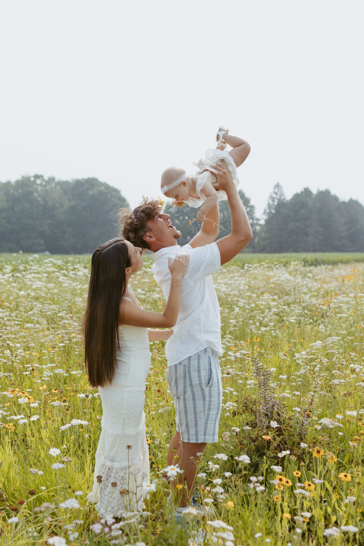 A young family stands in a wildflower meadow, father lifting baby in the air while mother watches joyfully.