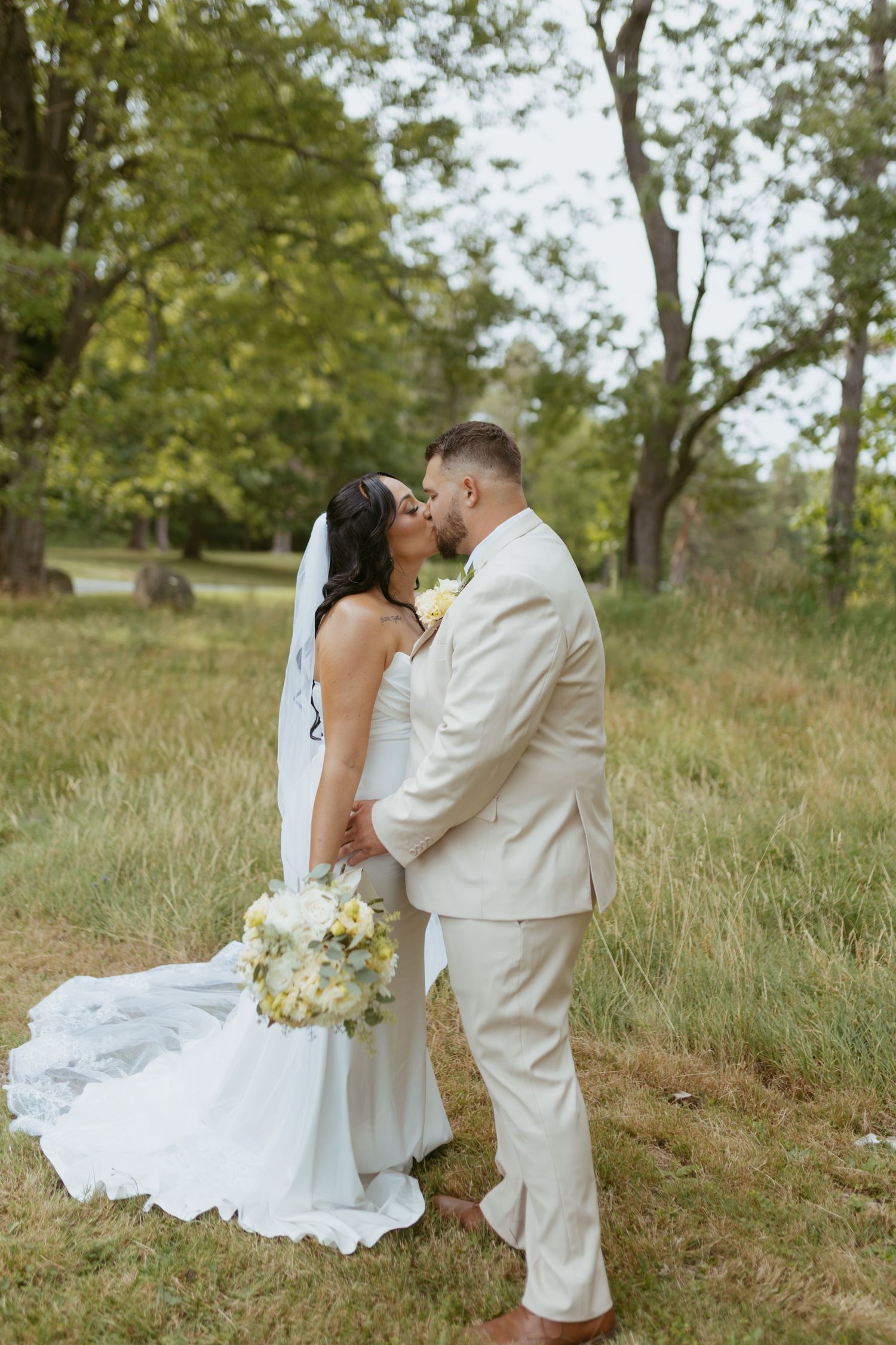 Bride and groom share a kiss in a lush green field, bride holding white bouquet in elegant gown with veil.