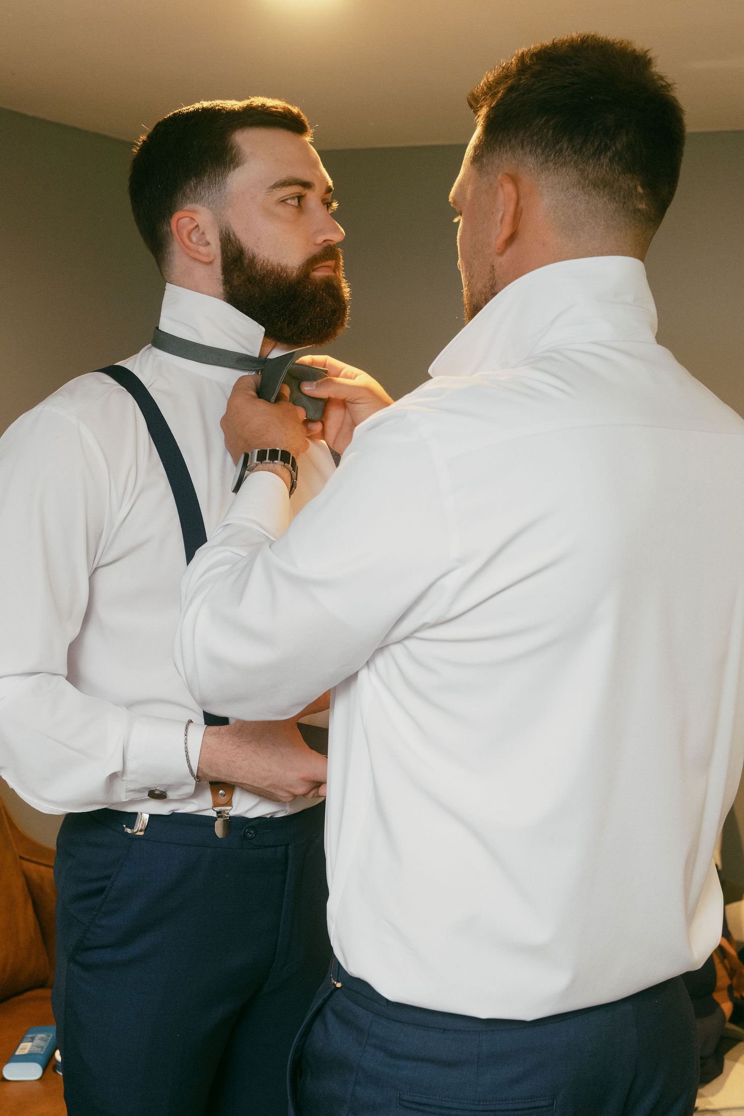 Groomsmen help adjust formal attire and suspenders during wedding preparation.