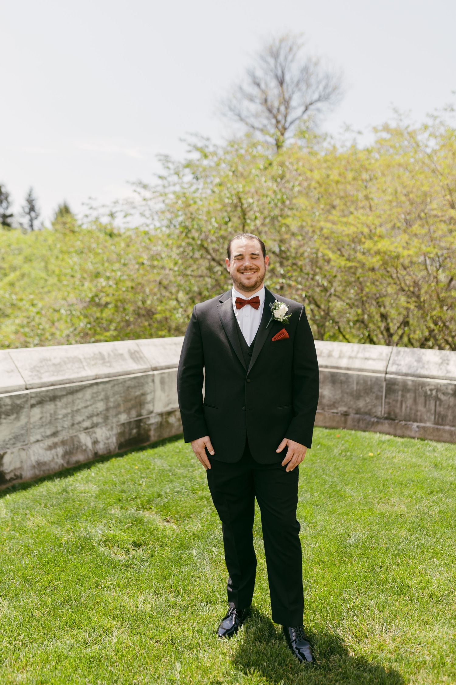 Groom in black tuxedo with red bow tie and boutonniere smiling outdoors on green lawn.