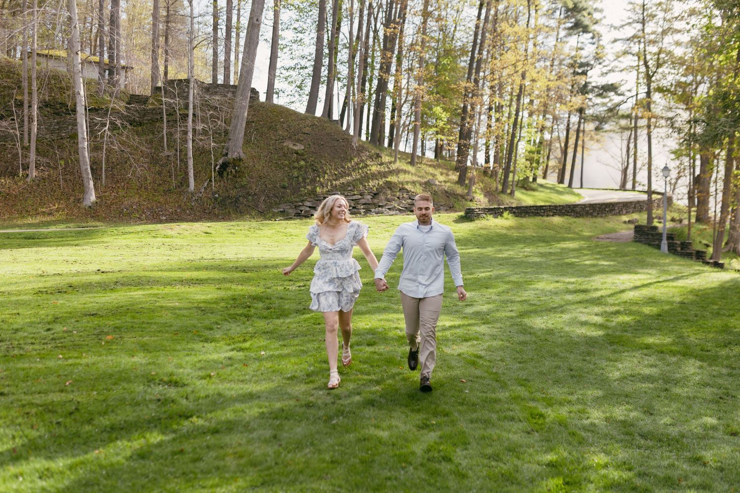 A couple holds hands and runs joyfully across a lush green lawn surrounded by tall trees in a scenic outdoor setting.