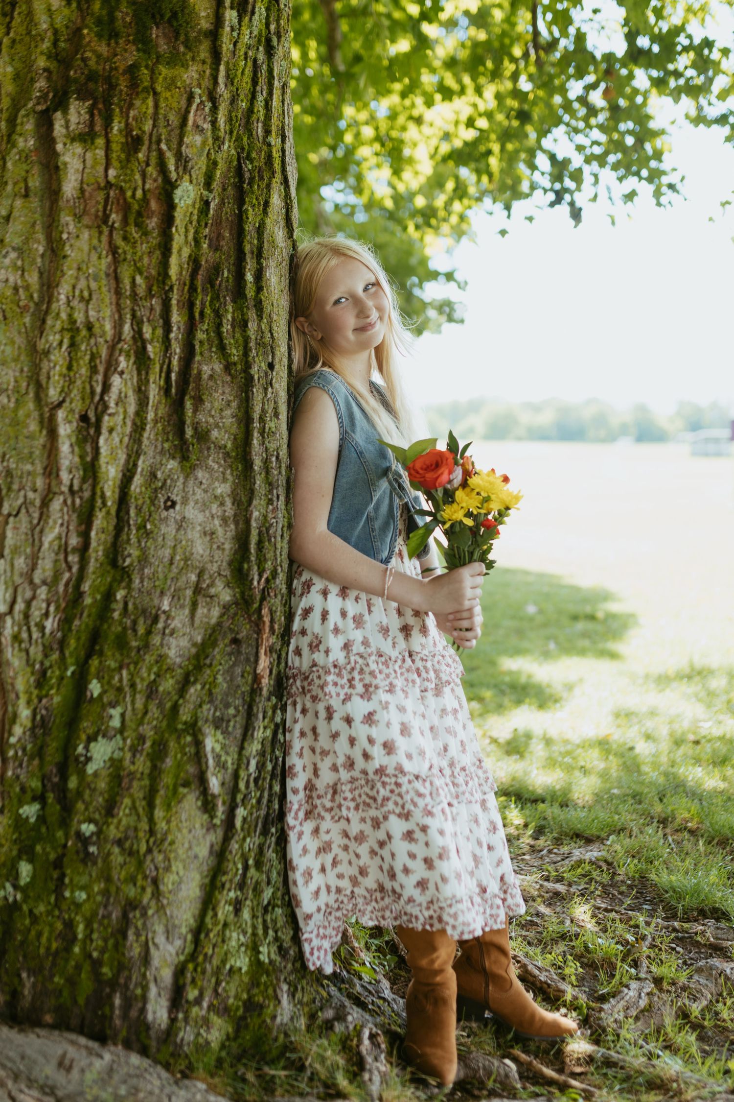 Young blonde woman in floral skirt and boots holding colorful flowers while leaning against a large tree outdoors.