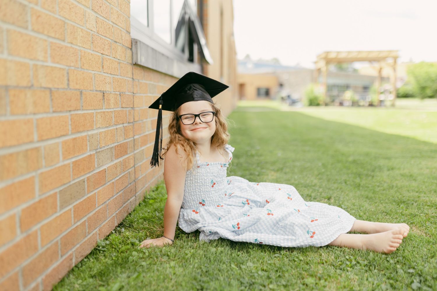 Young girl in graduation cap and glasses sits on grass against brick wall in floral dress, celebrating academic milestone.