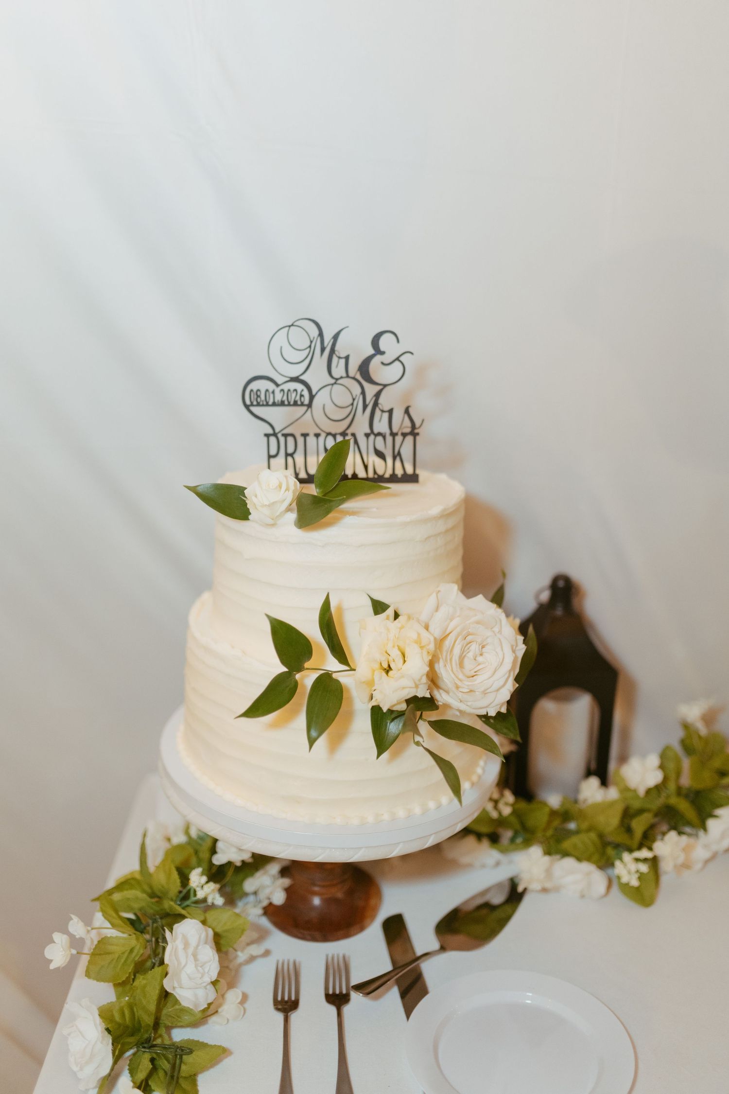 Two-tier white wedding cake with floral decor and Mr. & Mrs. Prusinski cake topper on elegant table setting.