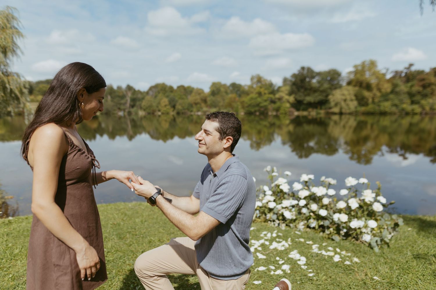 Man proposes to woman on one knee by a serene lake, holding a ring with white flowers nearby on a sunny day.