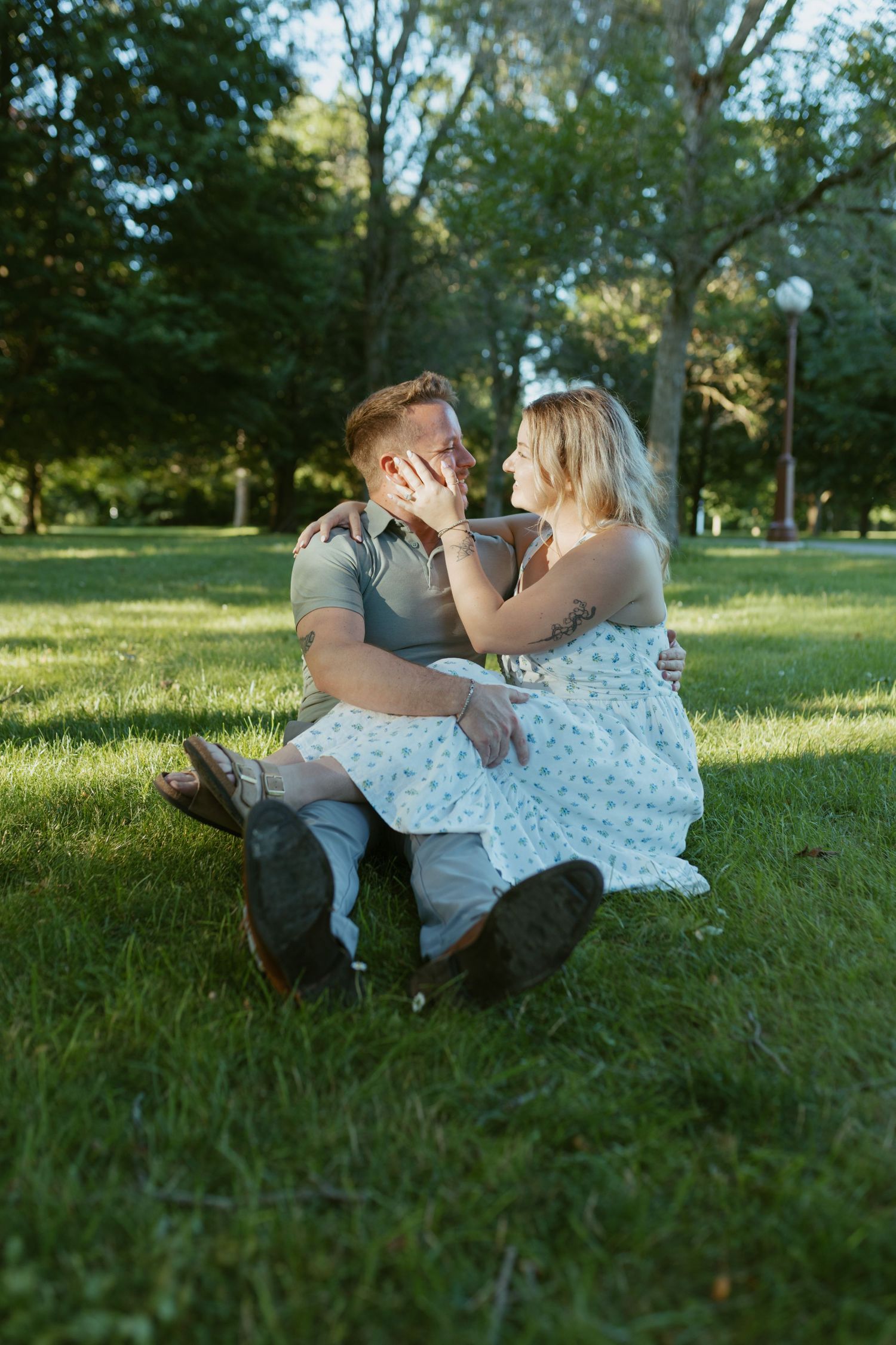 Couple sitting on green grass in a park, embracing and almost kissing, woman in floral dress, man in gray shirt.