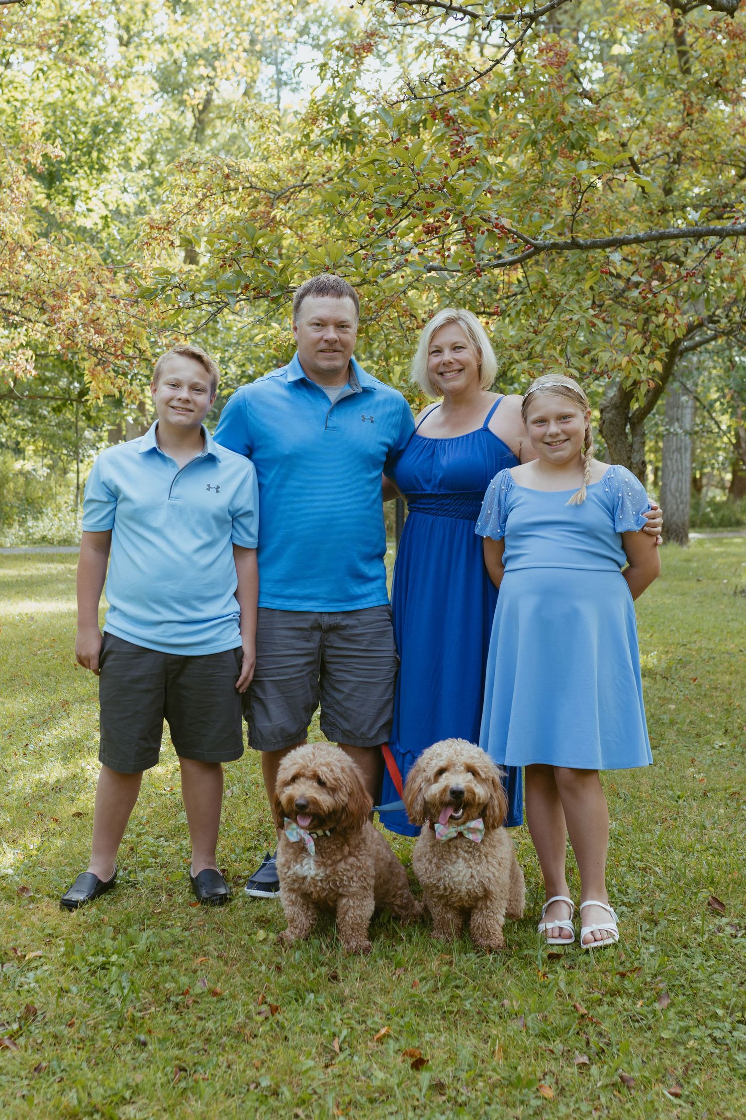 Family of four wearing blue clothing smiles outdoors with two goldendoodle dogs on a green lawn.