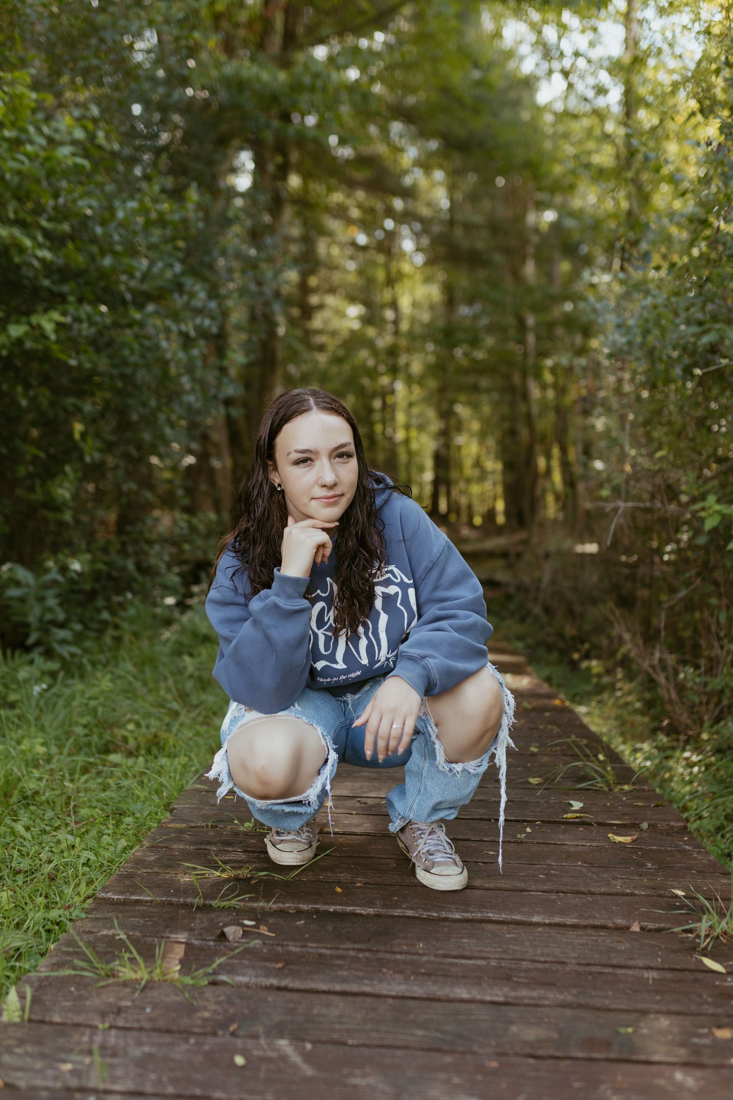 Young woman in blue hoodie and ripped jeans crouching on wooden path in lush green forest setting.
