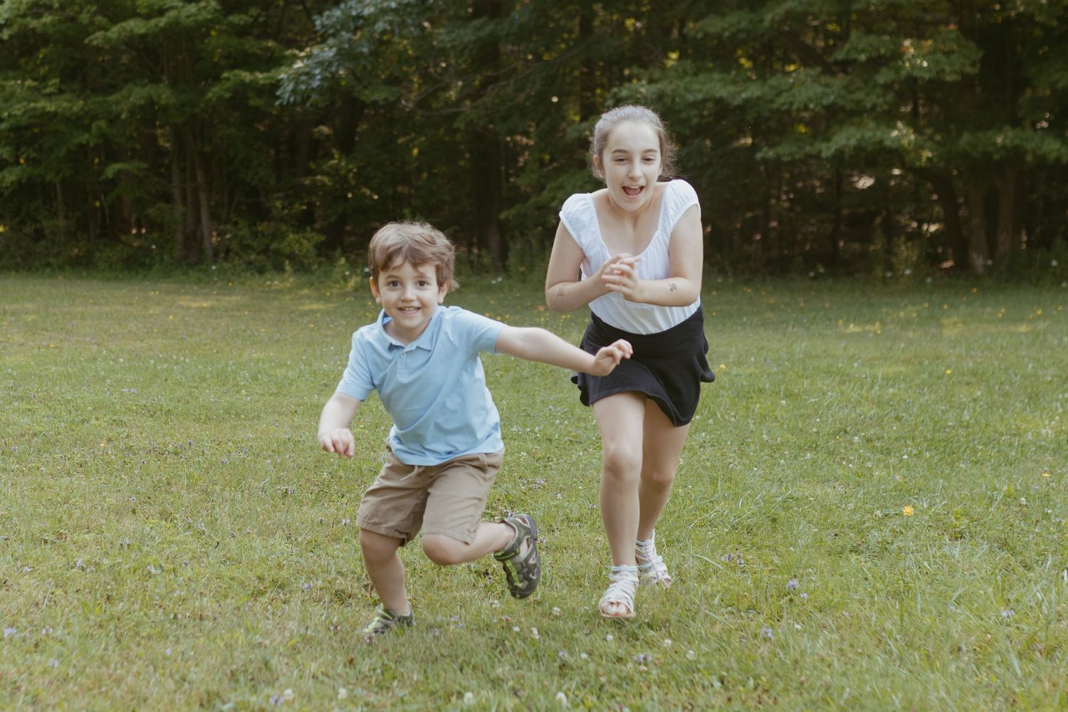Two happy children running and playing together on a green grass lawn with trees in the background.