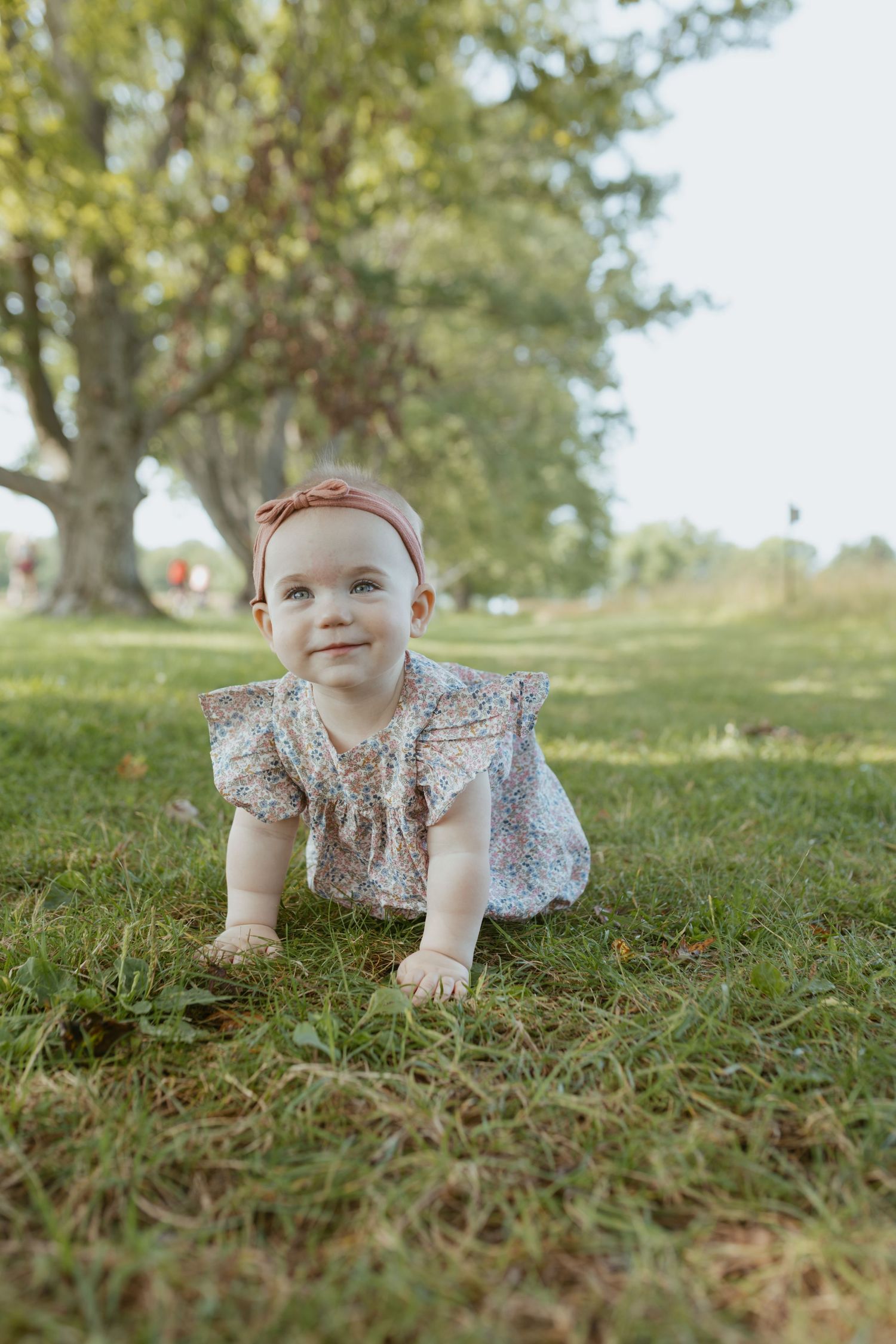 Baby girl in floral dress and pink headband crawling on green grass in a sunny park with large trees.