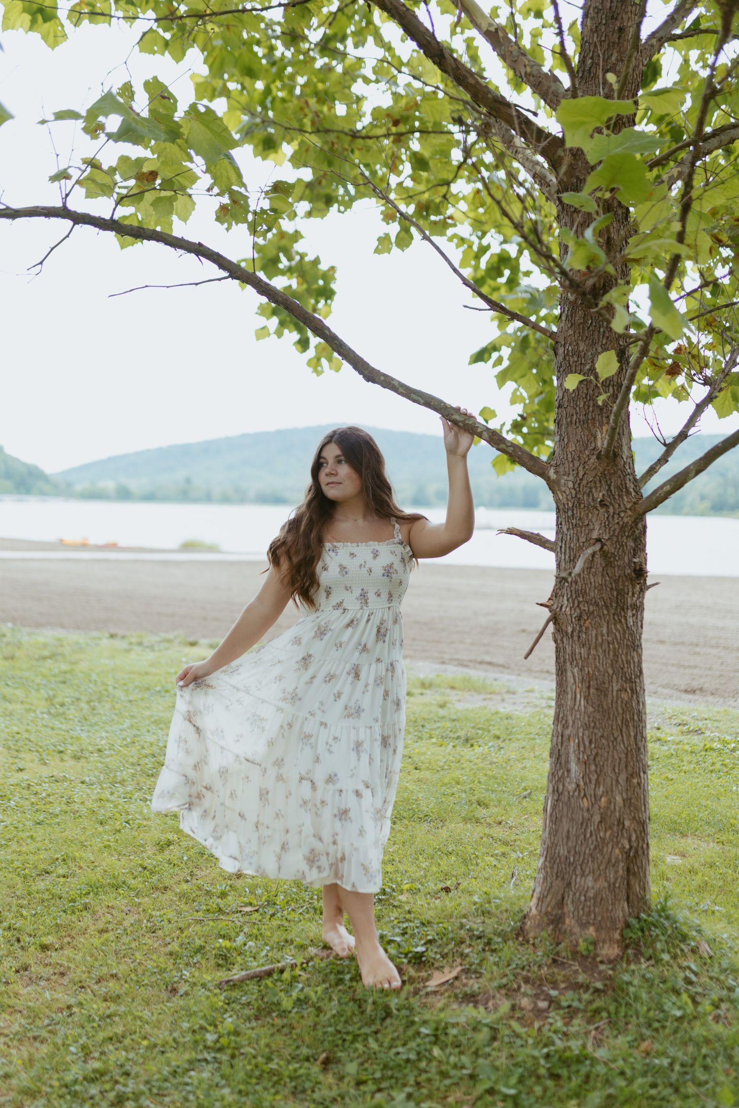 Woman in floral sundress standing barefoot by a tree near a lake shore with green hills in the background.