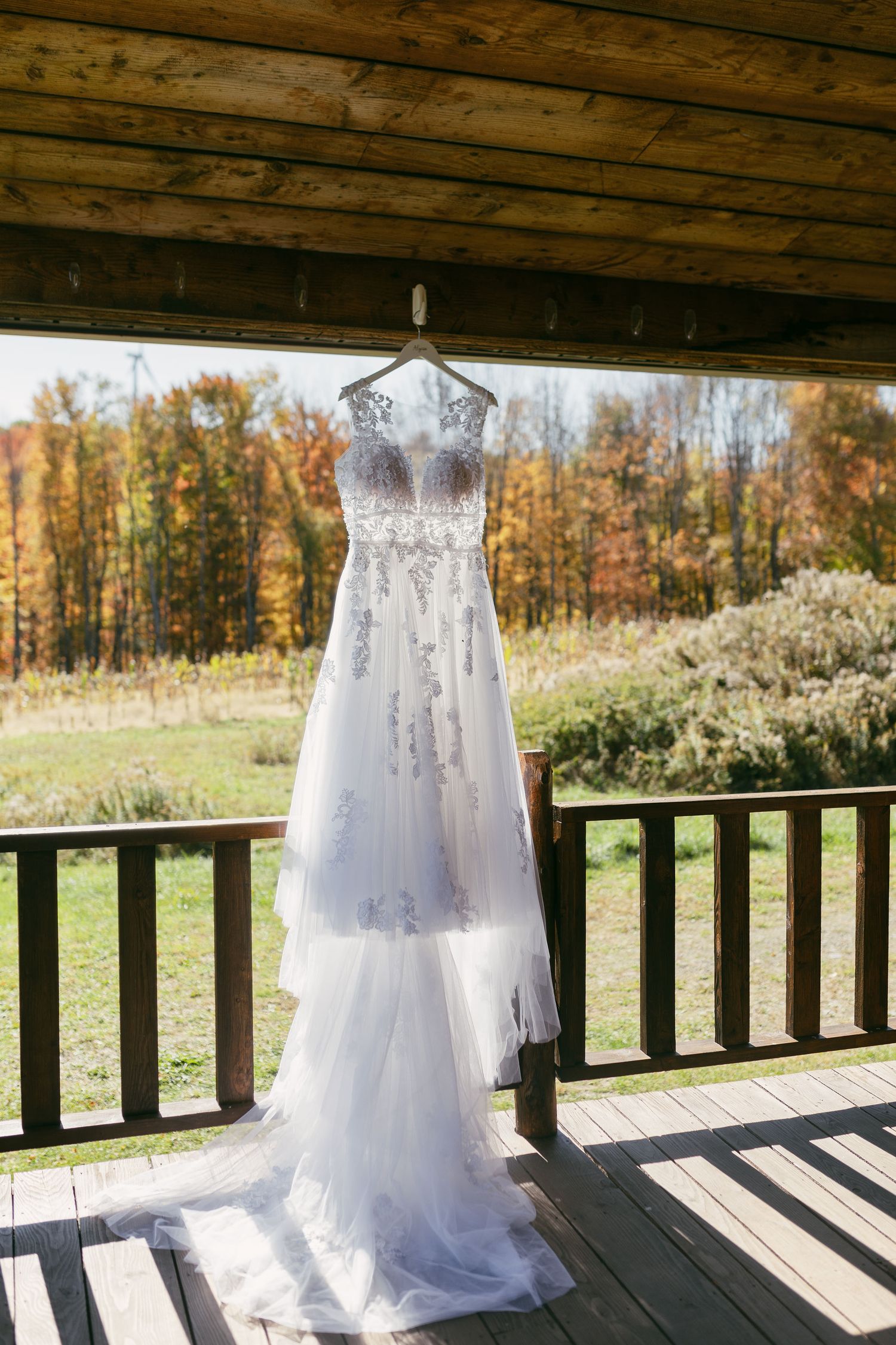 White wedding dress hangs on display along a rustic wooden porch railing with fall foliage in background.