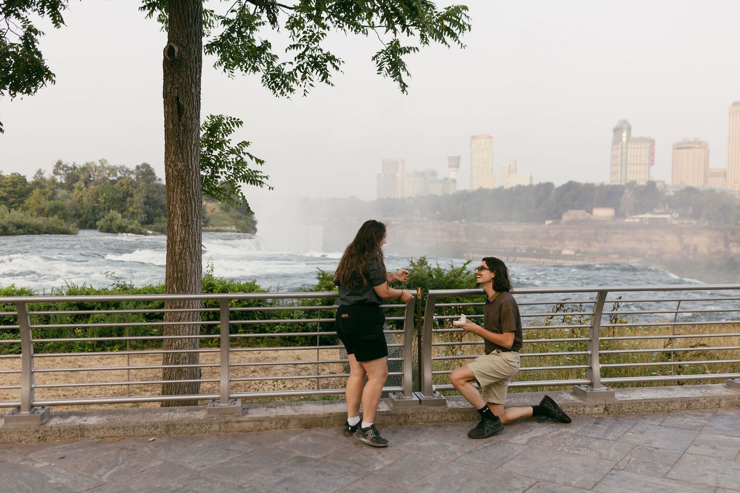Man proposing to woman on one knee at Niagara Falls overlook with city skyline in background.