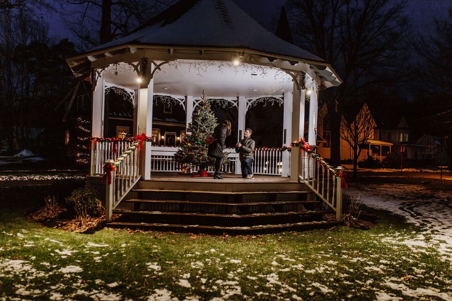 Couple sitting in a festively decorated gazebo at night with Christmas lights and snow-dusted ground.