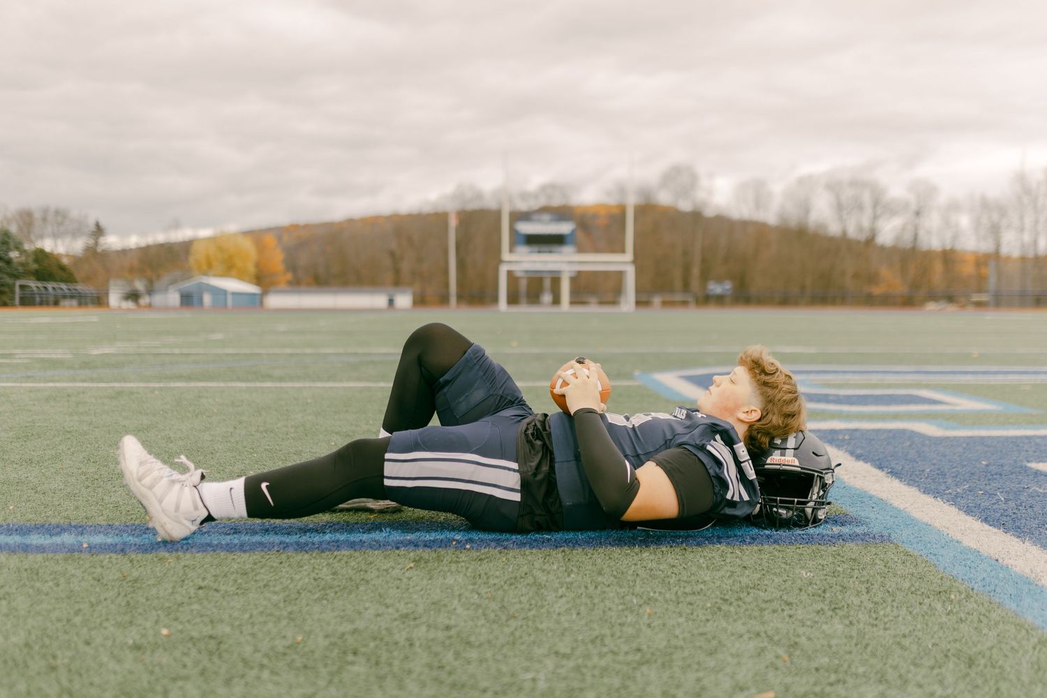 Football player lying on blue turf field holding helmet, wearing black uniform on cloudy autumn day.