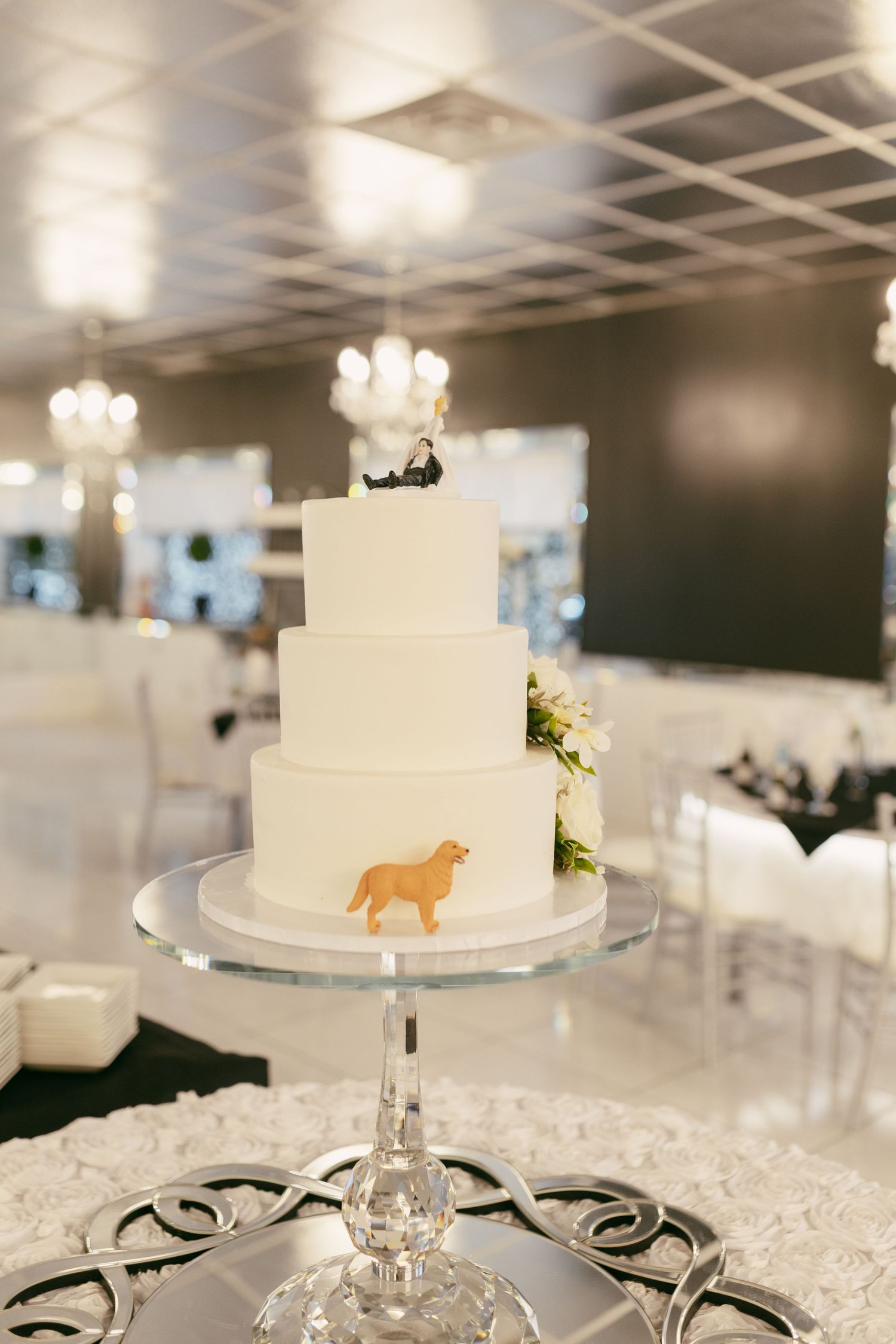 Elegant three-tier white wedding cake with golden dog silhouette decoration on crystal cake stand.