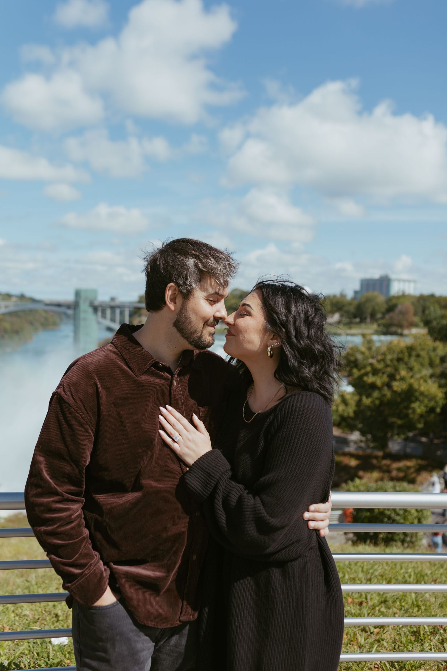 Couple sharing a romantic moment near Niagara Falls on a sunny day, woman in black dress, man in burgundy shirt.
