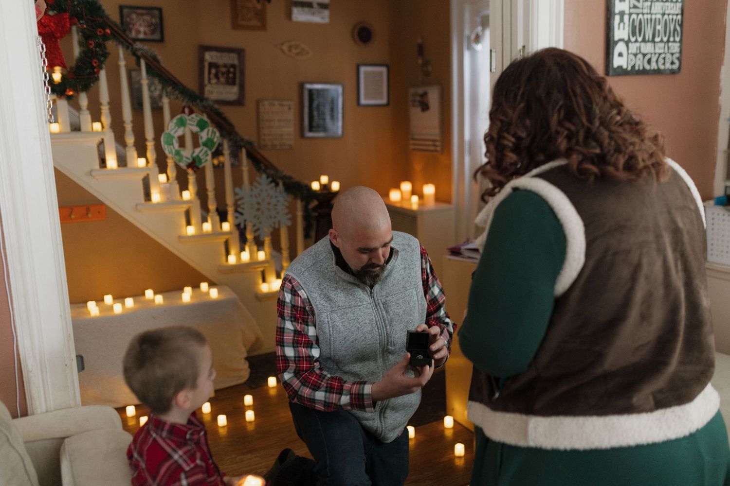 Man kneeling with ring box proposing to woman surrounded by candles on staircase with child watching holiday decor.