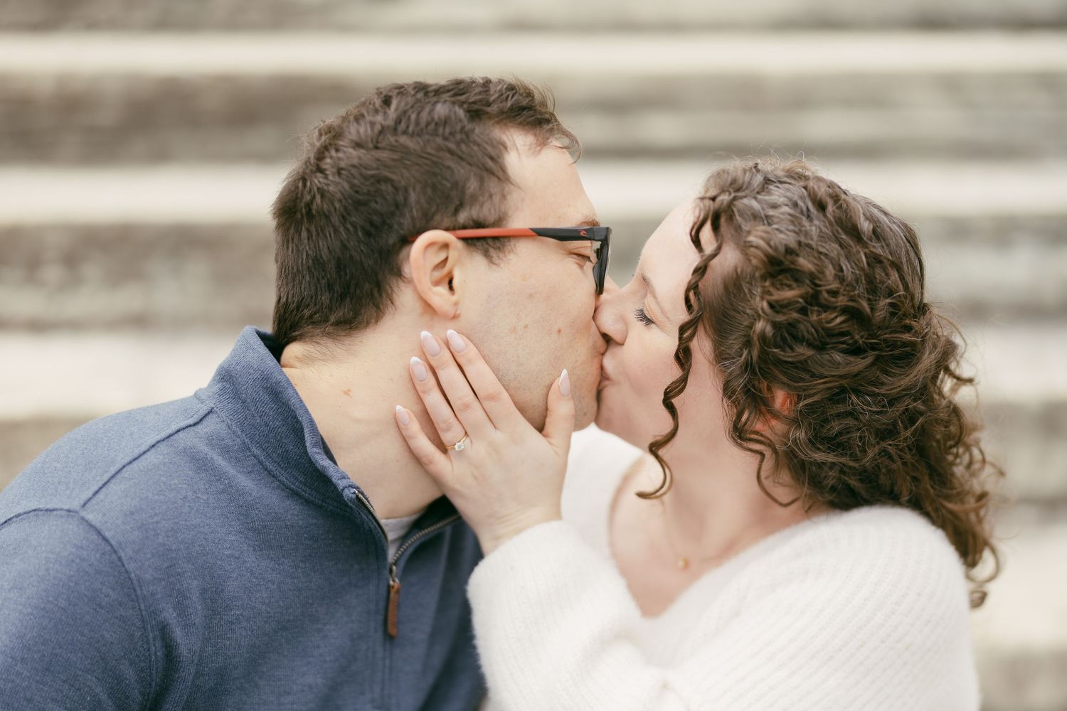 Couple sharing a romantic kiss on stone steps, man wearing glasses and blue sweater, woman in white sweater with curly hair.