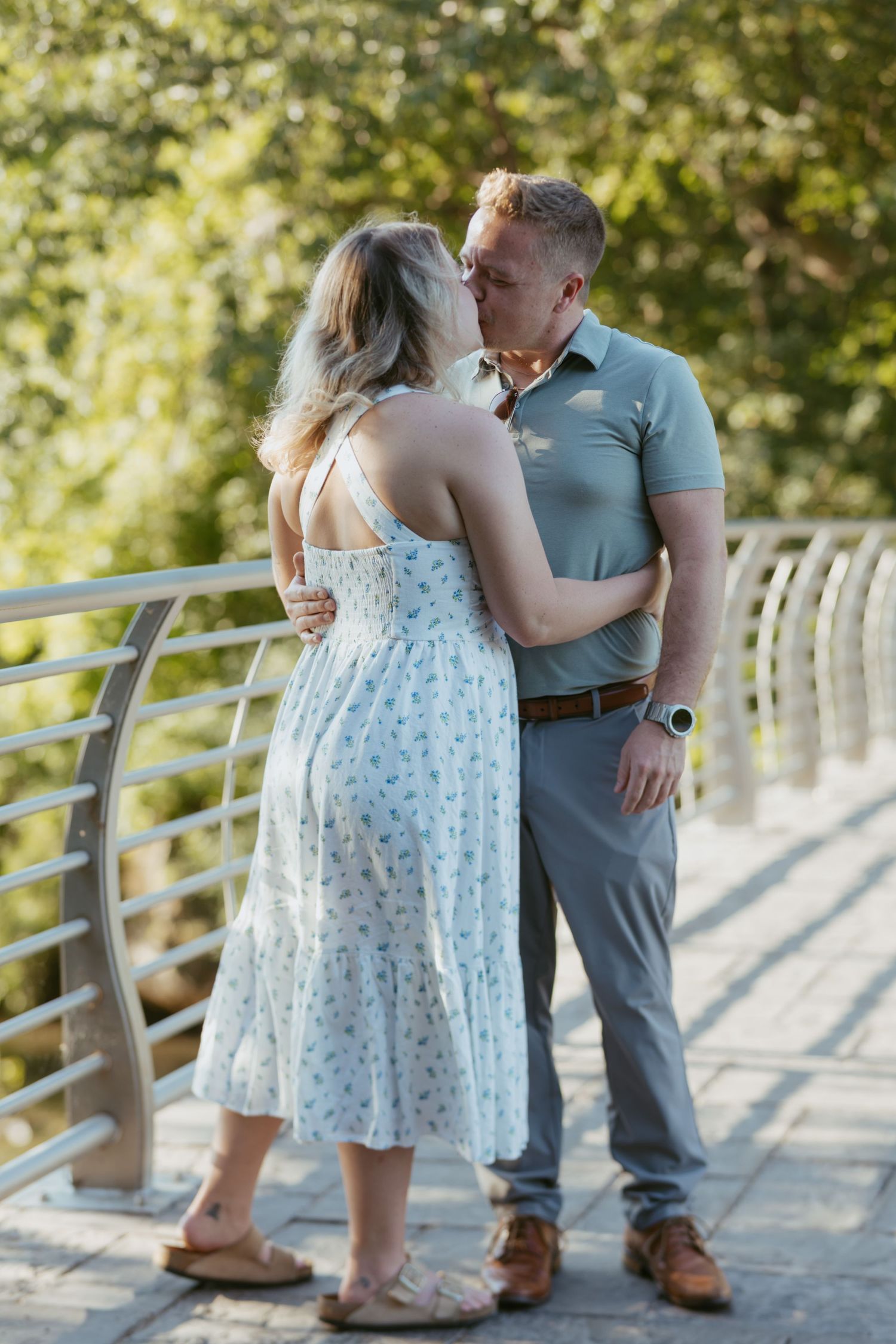 A couple shares a romantic kiss on a sunlit bridge, the woman in a floral dress and the man in a gray polo shirt.